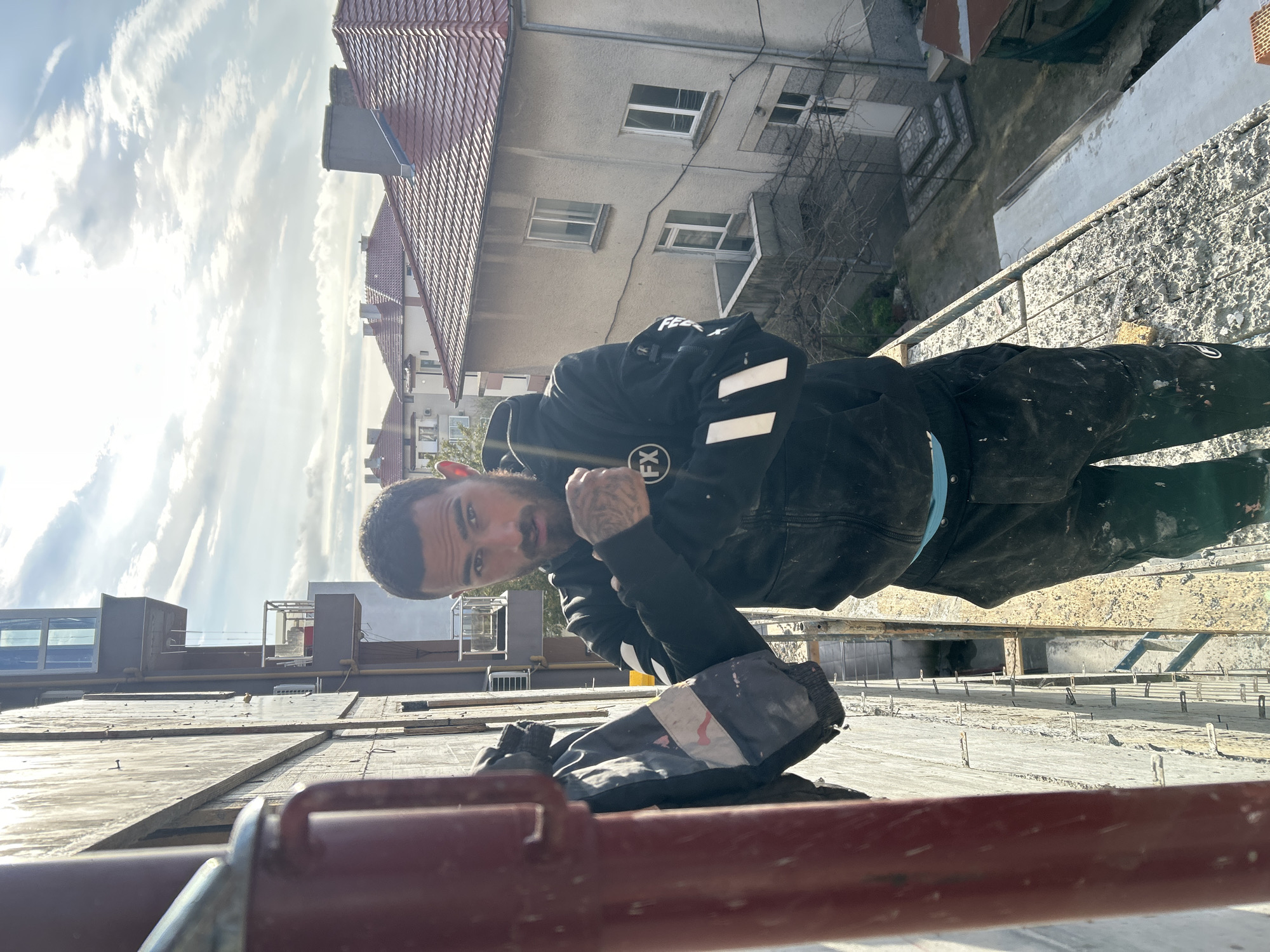 A man with a beard, tattoos, and dark clothing is photographed from below while standing on a structure in Tsarevo, Bulgaria. The man is looking down at the camera, with buildings and a cloudy sky in the background. The structure he is standing on appears to be under construction, with exposed rebar. A reddish-brown railing is in the foreground.