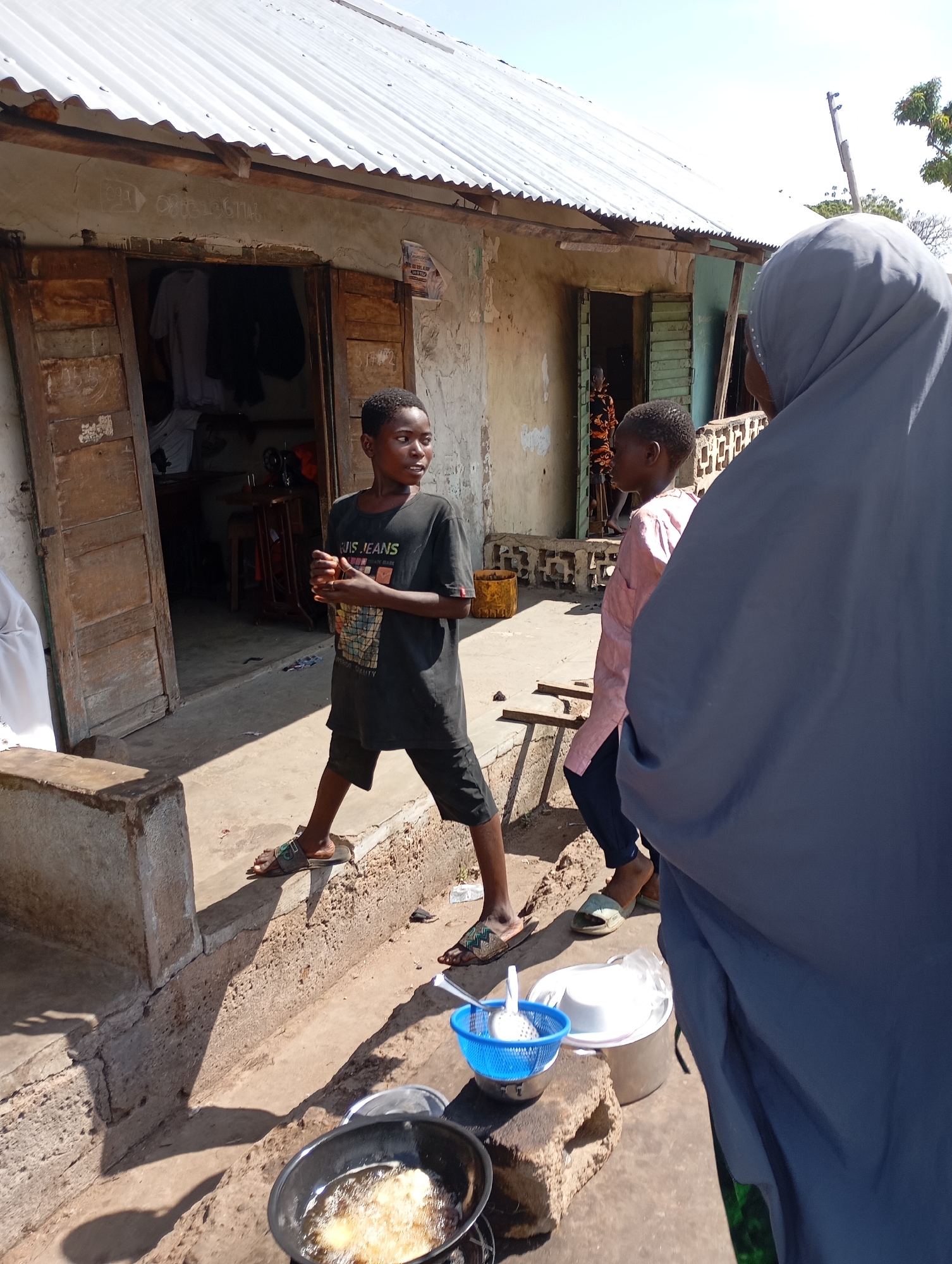 In Shendam, Nigeria, a young boy in a dark t-shirt and shorts stands near the entrance of a building with a corrugated metal roof. The building appears to be a shop or workshop, with wooden doors that are partially open, revealing clothing and a sewing machine inside. To the right of the boy, another child in a pink shirt and dark pants is visible, facing away from the camera. A person wearing a grey hijab stands in the foreground, obscuring part of the view. In front of the building, a cooking scene is set up on the ground, with a pot of oil on a fire, frying what appears to be food. Various bowls and utensils are also present, including a blue colander. The ground is made of concrete and dirt, with shadows cast by the building and people. The overall scene suggests a domestic or commercial setting in an outdoor environment.