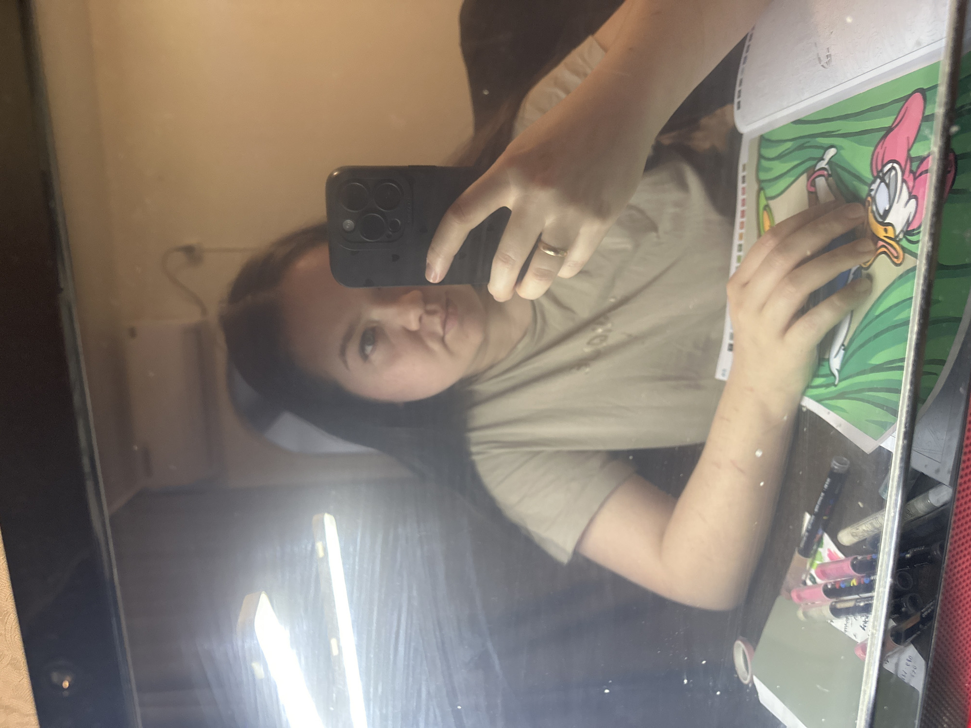 A selfie of a person with long dark hair, wearing a tan t-shirt and a gold ring on their finger, is reflected in a mirror. The person holds a black phone with three camera lenses in their left hand, covering their face partially. Reflected in the mirror is a wall outlet, a room with neutral-colored walls and a fluorescent light strip. On a surface to the right, there is a Daisy Duck drawing and multiple markers. Scars are visible on the person's left arm. It is impossible to confirm that this image was taken in the city of Chaikovsky, Russia.