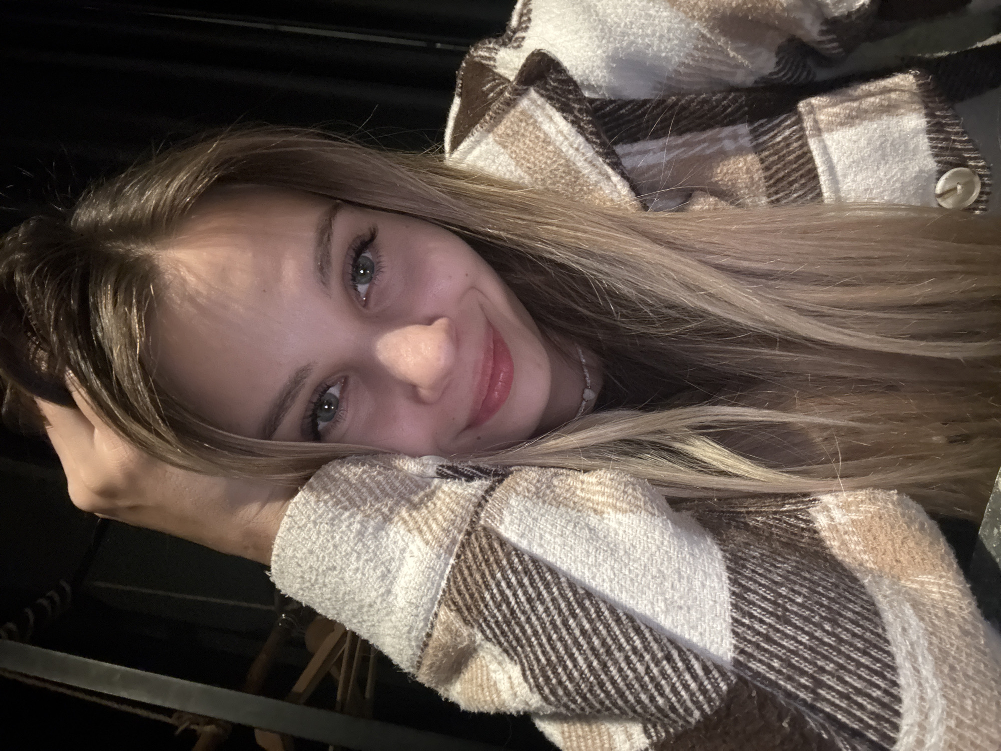 The image is a close-up, horizontally oriented selfie of a young woman with long, blonde hair. She is lying down, resting her head on her arm, which is mostly obscured by her plaid shirt. Her eyes are open and looking towards the camera, and she has a gentle smile on her face, with her lips slightly parted. She appears to be wearing light makeup, with subtle eyeliner and lip gloss. Her shirt is a cream-colored fabric with a brown and white plaid pattern, featuring a visible button on the right side. The background is dark and out of focus, with some hints of dark, possibly wooden, furniture or structure. The lighting suggests it might be evening or indoors. The overall mood is relaxed and warm, with the woman exuding a friendly and approachable demeanor. There is no visible text in the image. The location context is Chaikovsky, Russia.