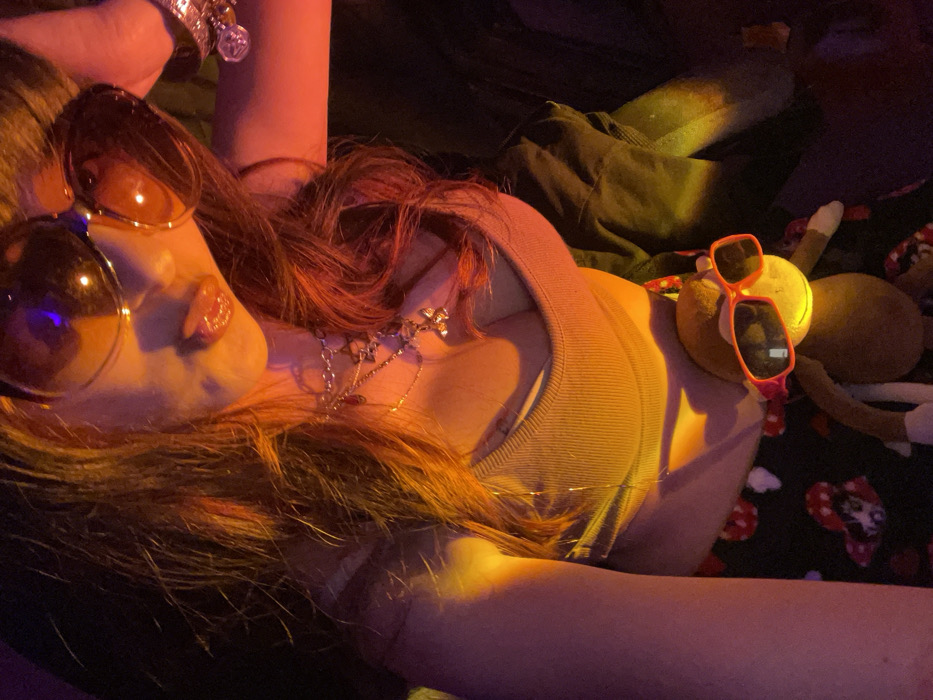 The image depicts a person, likely female, captured in a close-up, upward-angled selfie under prominent warm orange and yellow artificial lighting. The subject's upper face is obscured by large, dark, aviator-style sunglasses. Her lips appear glossy and slightly parted, revealing a glimpse of the tongue. Long, reddish-brown hair flows around her face and over her shoulders. She wears a light brown ribbed crop top, multiple layered necklaces with silver chains and charms, including a butterfly and a red droplet, and a charm bracelet on her right wrist. Her lower garment is dark with a white and red mushroom pattern. A brown plush monkey toy, adorned with small orange-red sunglasses, rests in the lower center of the frame, positioned in front of the subject. The background is dark and indistinct, suggesting an indoor environment. The image contains no identifiable visual cues to determine a specific location within Raanana, Israel.