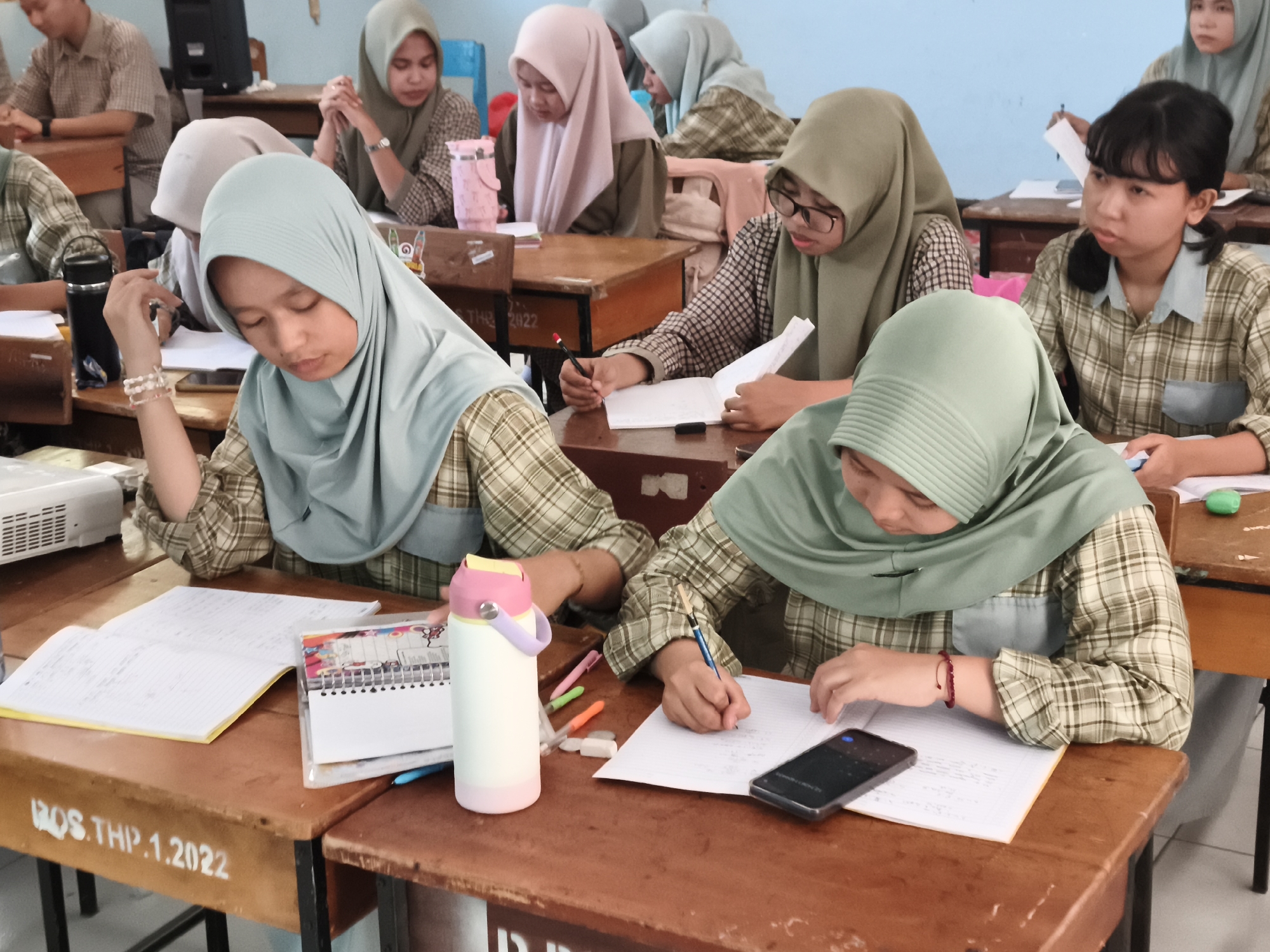 The image shows a classroom in Makassar, Indonesia, where several students are seated at wooden desks, engaged in what appears to be a learning activity. The students are predominantly female and are wearing hijabs, with most of them in plaid school uniforms.
In the foreground, two female students are the primary focus. The student on the left, wearing a light blue hijab, is looking down at a notebook, with her right hand resting on the paper. She is wearing a beaded bracelet on her wrist. Next to her is a black thermal mug and a spiral-bound notebook.
The student on the right, also wearing a light blue hijab and a plaid shirt, is actively writing in a notebook with a pencil. A smartphone is placed on her notebook, and a red beaded bracelet is visible on her wrist.
In the background, other students are also visible. One student in a pink hijab is looking down at her work. Another student in a dark green hijab is wearing glasses and is also focused on her notebook. Further in the background, a teacher or another student in a light brown plaid shirt is visible. The classroom environment is characterized by wooden desks and chairs, with white walls and a light blue section visible in the upper right corner. The desks have handwritten markings, including "BOS.THP.1.2022" on one of them. The overall atmosphere suggests a typical school day with students concentrating on their studies.