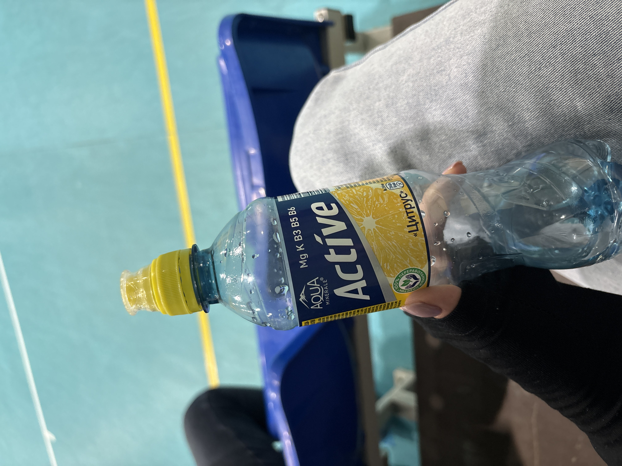 The image shows a close-up of a person holding a clear plastic bottle of water in what appears to be an indoor sports facility in Votkinsk, Russia. The person's lap, clad in light grey denim-like fabric, and a hand wearing a black fingerless glove or sleeve with light pink nails are visible.
The bottle has a bright yellow cap and a yellow and blue label. Condensation on its surface suggests the drink is cold. The label clearly displays the brand "AQUA MINERALE® Active" and highlights the inclusion of "Mg K B3 B5 B6" (Magnesium, Potassium, and B vitamins). The flavor is indicated in Cyrillic as "Цитрус" (Citrus), accompanied by a graphic of a citrus fruit slice. A notable detail on the label is the text "100% ПЕРЕРАБОТАННЫЙ ПЛАСТИК," which translates to "100% RECYCLED PLASTIC," along with a recycling symbol.
The background reveals a light blue floor with distinct yellow and white lines, typical of a sports court or gymnasium. Part of a blue plastic seat or bench is also visible. The setting suggests an athletic event, practice, or spectating environment. The image is taken from a slightly downward angle, focusing on the bottle and the person's hand. The activity is limited to holding the bottle, implying a moment of rest or hydration. The scene is well-lit, indicating an indoor environment.