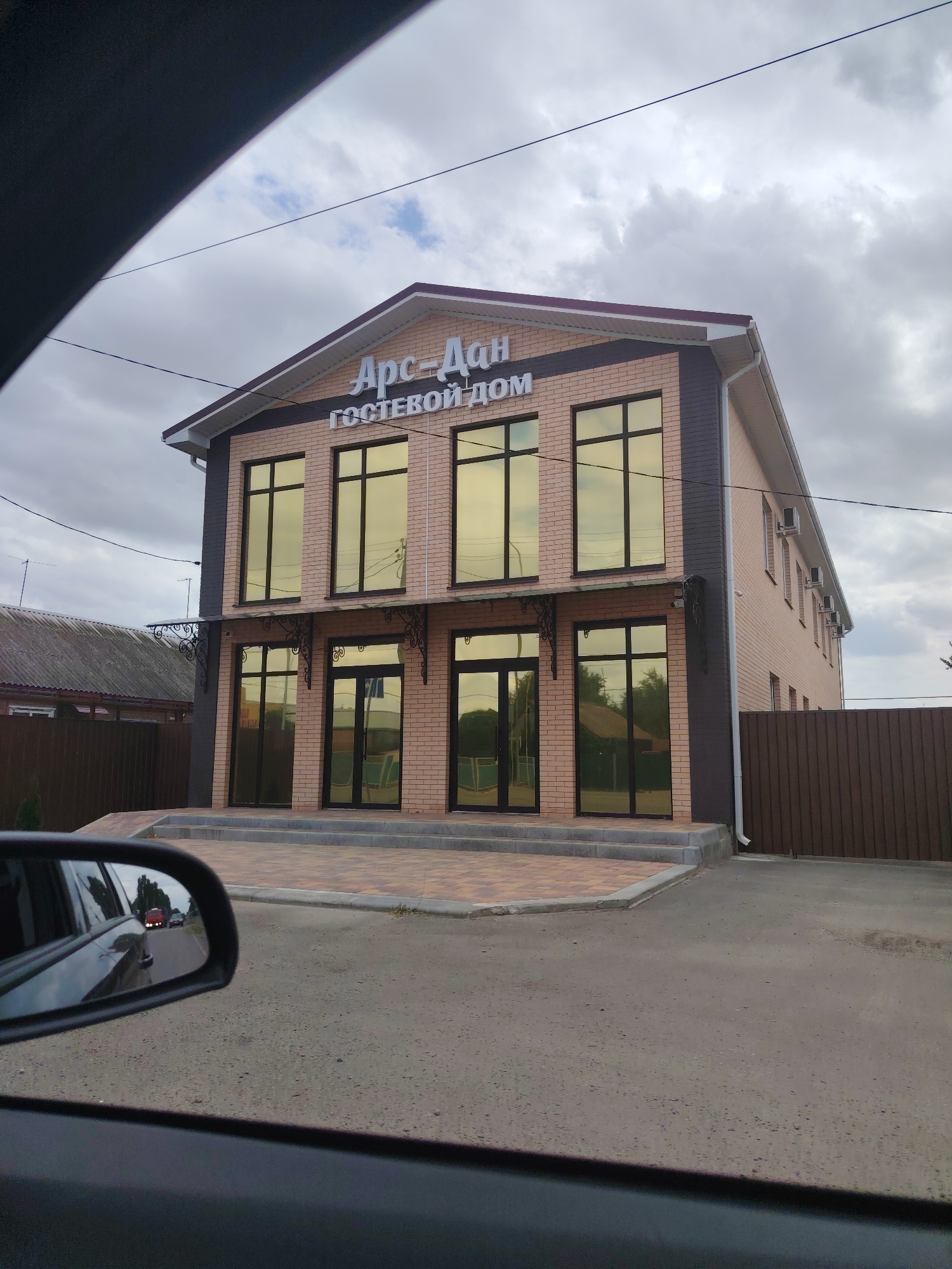 The image shows a two-story brick building with large golden-tinted windows, identified by the signage "Арс-Дан Гостевой Дом" as "Ars-Dan Guest House". The building is located on a paved street in the city of Tselina, Russia. The facade of the building is a light brick color with darker brick accents on the corners and around the windows. There are two entrances on the ground floor, both with double glass doors. Above the entrances, decorative metal brackets support small awnings. The building has an air conditioning unit mounted on the wall to the right. A brown metal fence runs along the right side of the property, and a smaller, older-looking building with a corrugated roof is visible to the left. The sky is overcast with gray clouds. In the foreground, a car's side mirror reflects a road with other vehicles. The overall scene depicts a commercial establishment in a small Russian town.