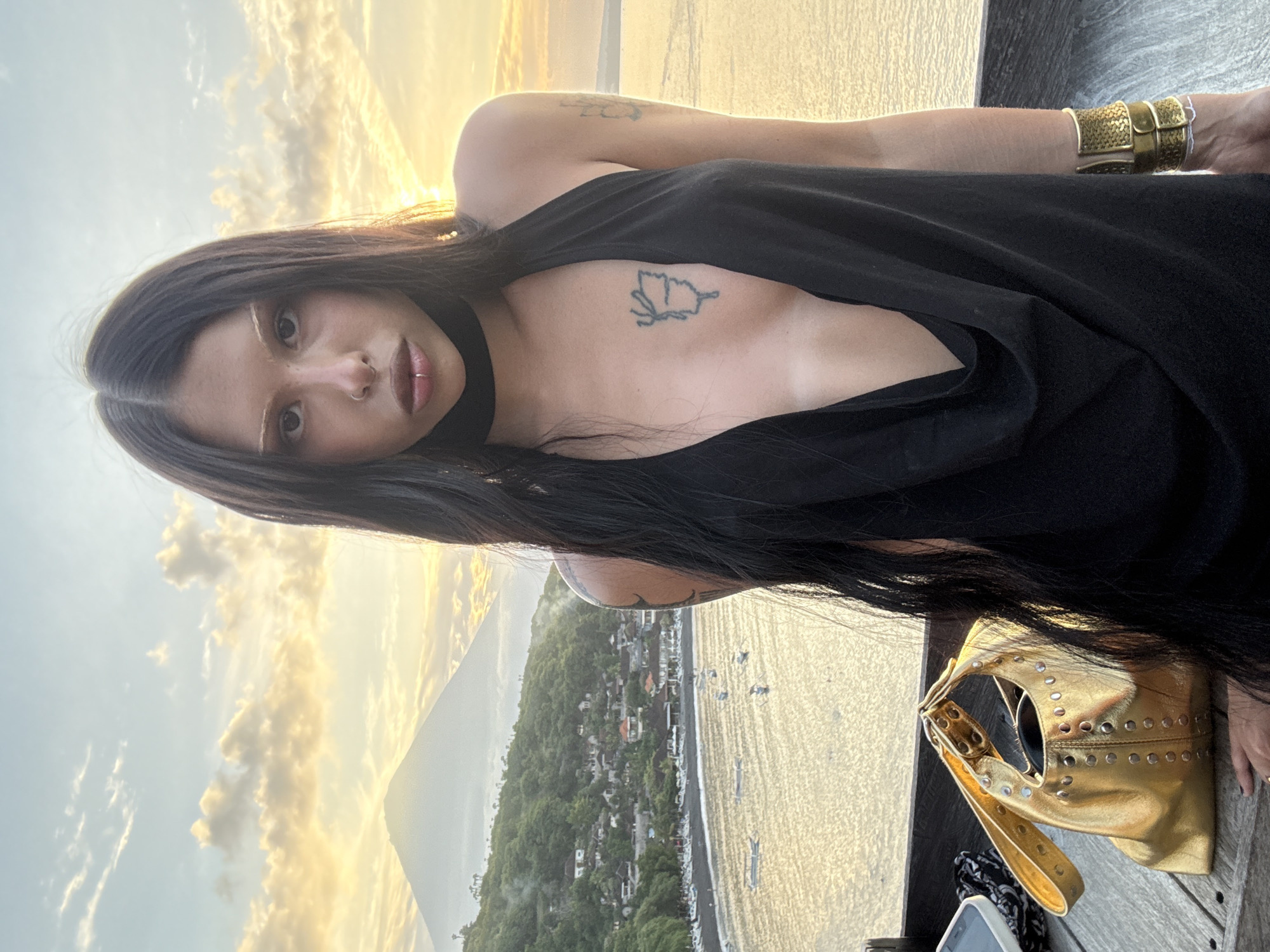 A young woman with long, dark hair and light-colored eyebrows is posing for a portrait against a sunset backdrop. She is wearing a black, sleeveless dress with a deep V-neckline that reveals a butterfly tattoo on her chest. She has lip and nose piercings, and a choker necklace. A metallic gold handbag sits on the wooden surface in front of her. In the background, a lush green hillside with houses overlooks a body of water with boats. A mountain rises in the distance, bathed in the warm glow of the setting sun. The overall scene is one of tropical tranquility and personal style.