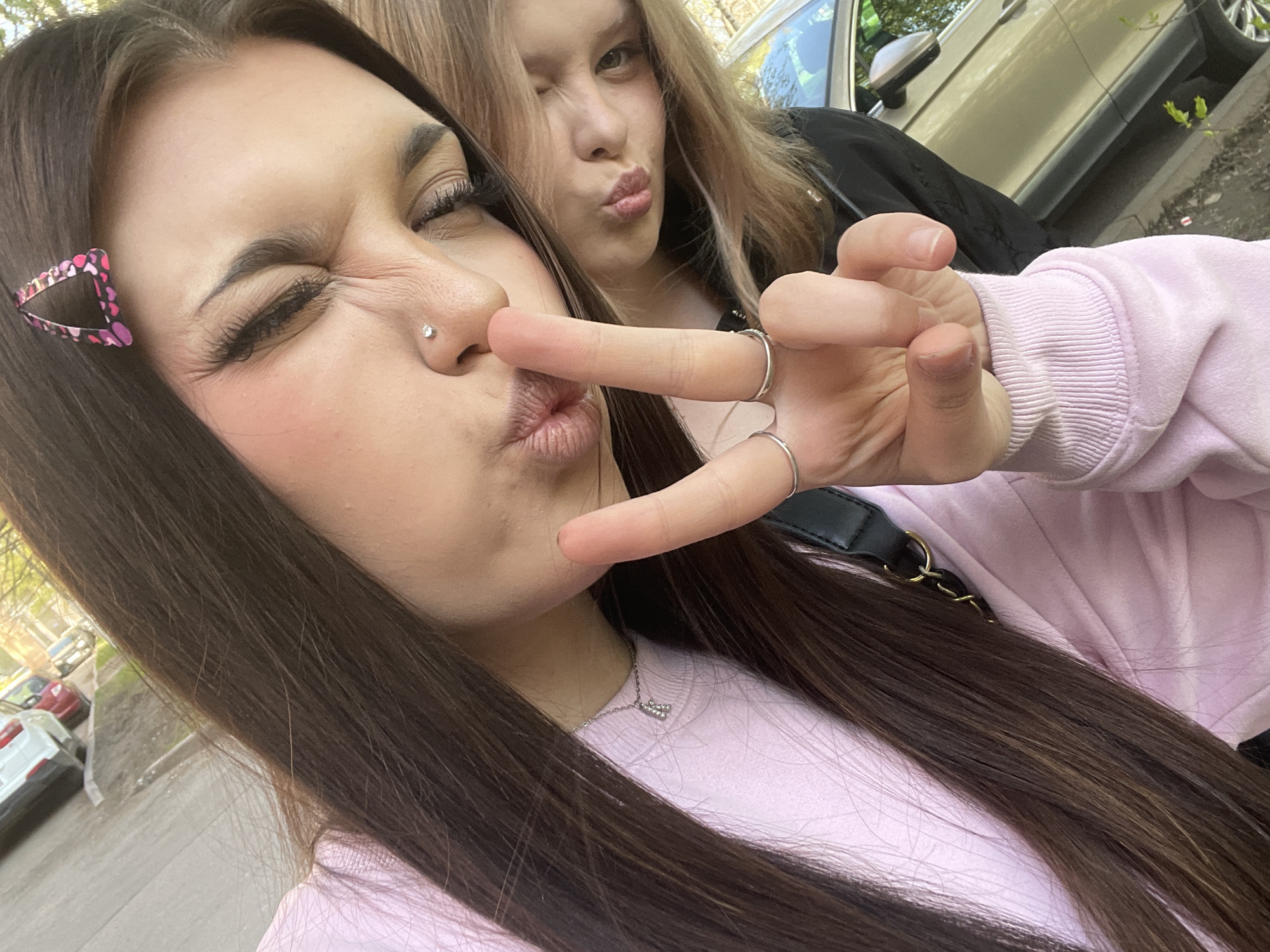 Two young women pose for a selfie outdoors. The woman in the foreground, with long dark hair and a pink triangle hair clip, wears a light pink sweatshirt and a silver necklace. She has a nose piercing and several rings on her fingers. She puckers her lips and winks one eye while holding two fingers up. The second woman stands behind her, also puckering her lips and winking one eye. She has shoulder-length blonde hair and wears a black jacket. Behind them, a light-colored SUV is visible, parked on a street. Other cars are parked in the background. The setting appears to be an urban environment, possibly a city street.