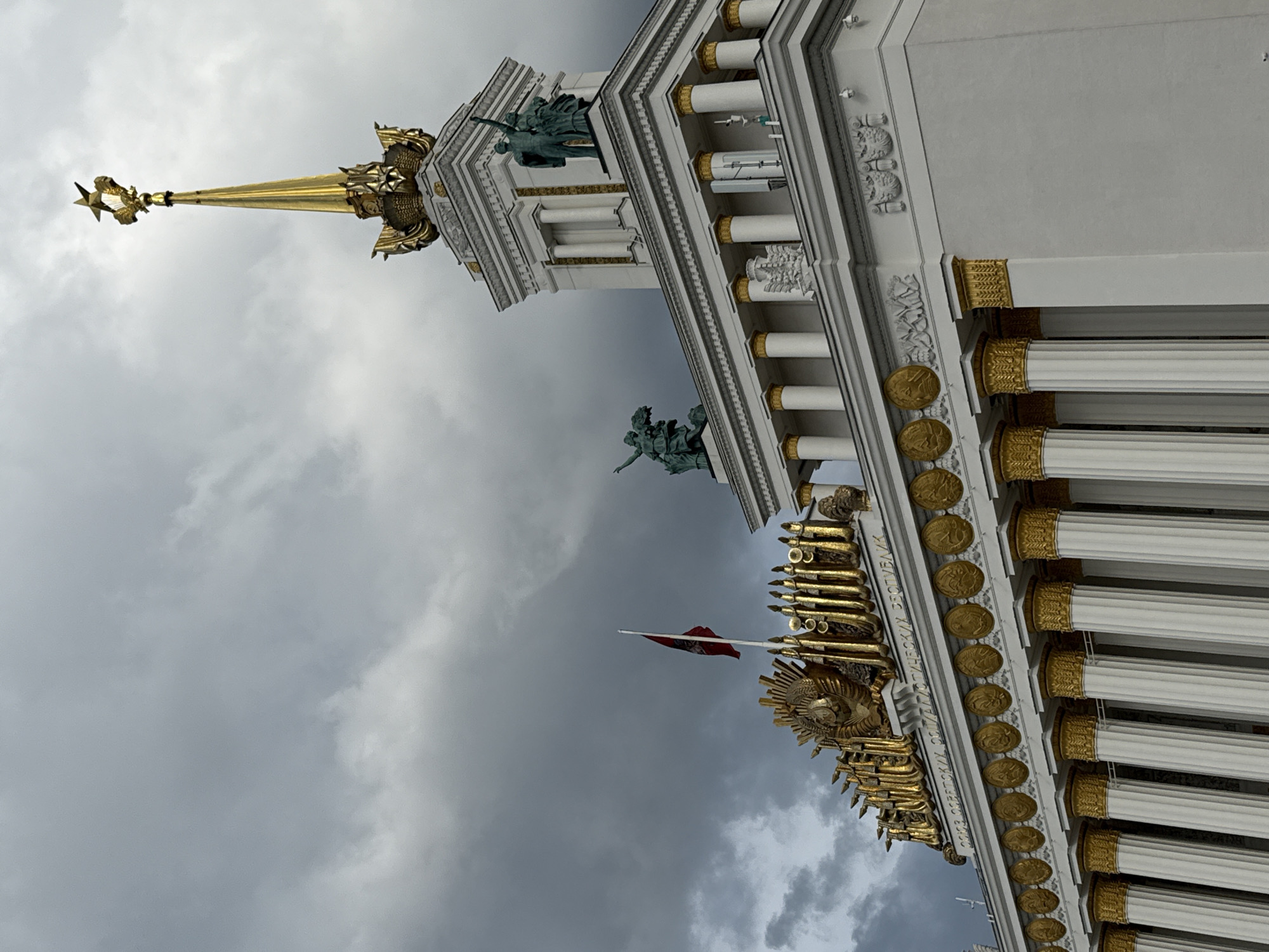 This low-angle, vertical shot captures the ornate facade of a grand building, likely an exhibition hall or government structure, against a cloudy sky. The architecture is predominantly white with striking gold accents. Prominent features include rows of fluted columns topped with gold capitals, a decorative frieze, and circular medallions depicting profile busts.
A towering spire, also golden, culminates in a five-pointed star, suggesting a Soviet-era landmark. Atop the spire and along the building's upper levels are sculptural figures, appearing to be made of a green-patinated bronze or similar material. One statue on the right depicts a figure reaching upwards. Further down the facade, a red flag with a crest waves from a flagpole. Below the flag, the building is adorned with a large, golden emblem resembling a sunburst or star, with what appears to be a Soviet coat of arms or similar symbol in the center.
Text is visible on the building, though partially obscured and in Cyrillic script. The legible parts appear to read "СОЮЗ СОВЕТСКИХ СОЦИАЛИСТИЧЕСКИХ РЕСПУБЛИК" (Union of Soviet Socialist Republics) and "ДРУЖБА НАРОДОВ" (Friendship of Peoples).
The weather is overcast with dramatic grey clouds. The lighting suggests it is daytime, possibly late afternoon or early morning given the diffused light. The overall impression is one of monumental grandeur and historical significance. The building itself is the primary subject, with the sky and architectural details contributing to the scene's setting in Moscow, Russia. No specific activity or event is depicted beyond the static presence of the building and its adornments.