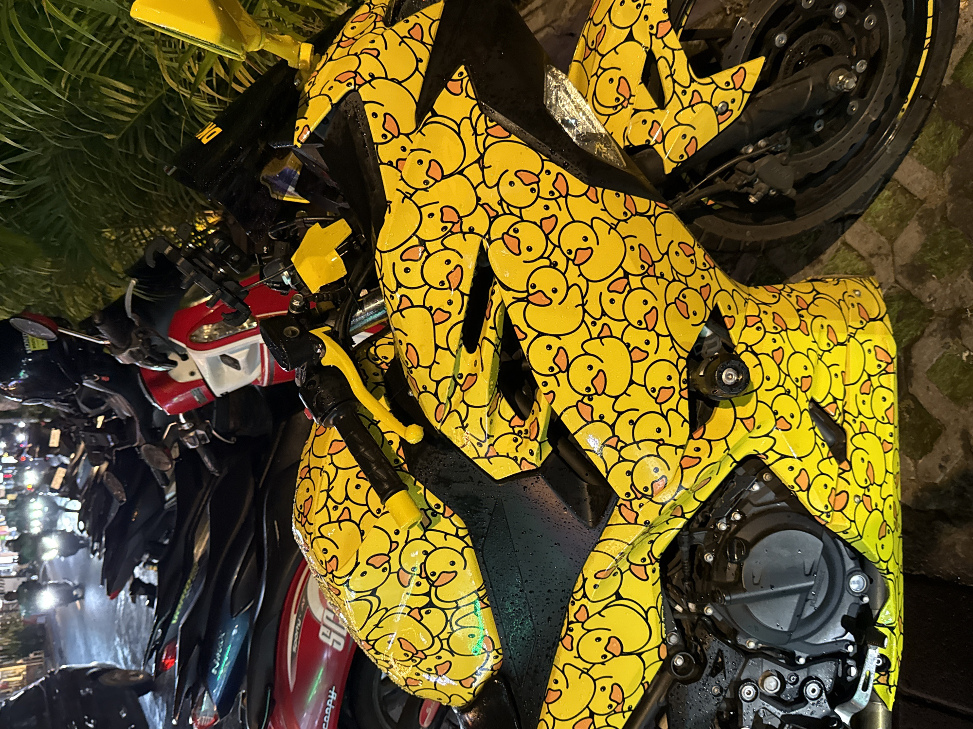 The image shows a motorcycle parked on a street in North Kuta, Indonesia. The motorcycle is custom-painted with a yellow background and numerous rubber ducky cartoon drawings on its panels. Other motorcycles are parked next to it, and some blurred figures can be seen in the background near a light source. The motorcycle appears to be wet, with droplets visible on its surface.