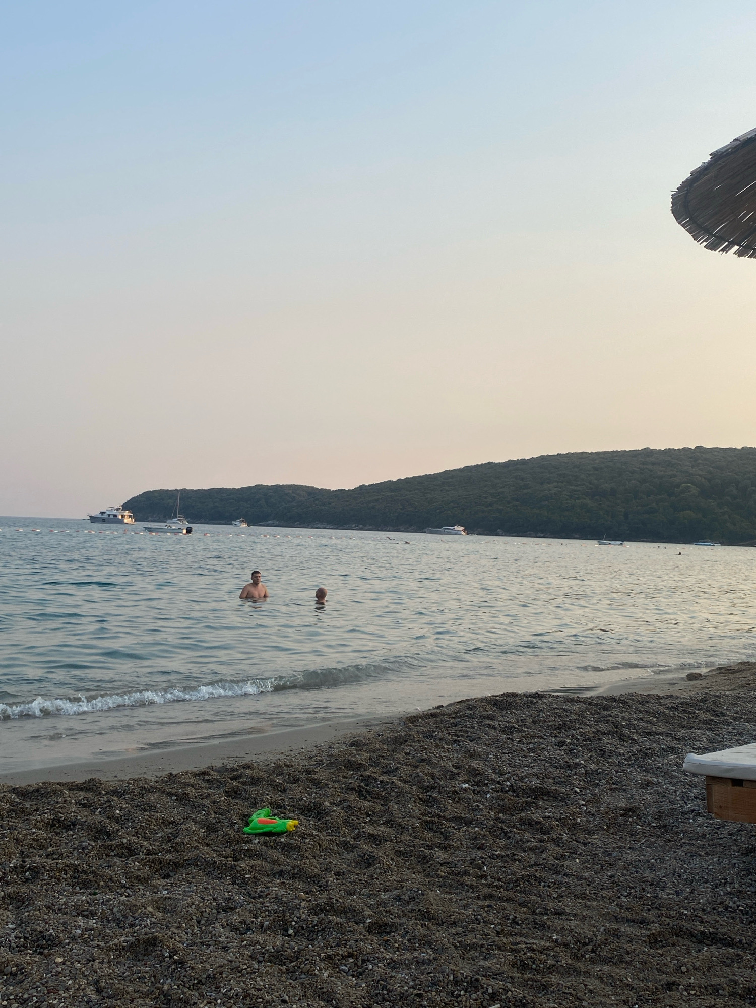 The image shows a serene beach scene in Prijevor, Montenegro. The water is calm and reflects the soft light of what appears to be late afternoon or early morning. Two men are visible wading in the shallow water, their bodies partially submerged. In the background, several boats are anchored or cruising on the sea, and a lush, tree-covered hillside forms the backdrop. The foreground is a beach covered in dark pebbles, with a small, colorful toy visible on the sand. A portion of a straw umbrella can be seen in the upper right corner of the frame, suggesting a lounging area. The sky is clear and graduates from a pale blue at the top to a warmer, lighter hue near the horizon.