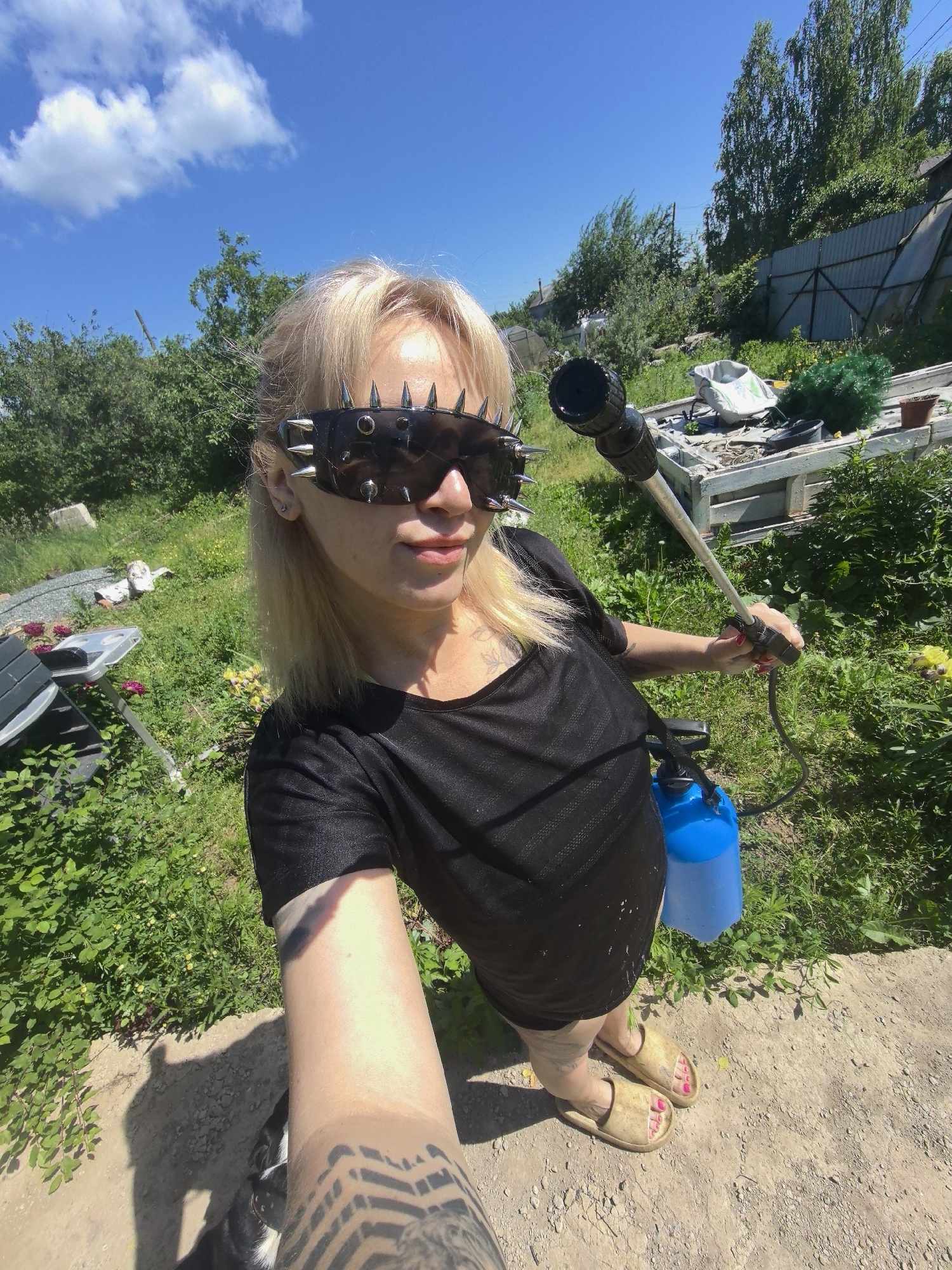 A fair-skinned woman with blonde hair, wearing a black t-shirt and studded sunglasses, stands in a yard in Noviy, Russia, holding a spray bottle and hose. She is looking into the camera. The yard is overgrown with grass and trees, and a metal fence can be seen in the background. The sky is blue with some clouds. A dog is partially visible in the lower left corner of the image. The woman has tattoos on her arm. She is wearing tan slides on her feet.
