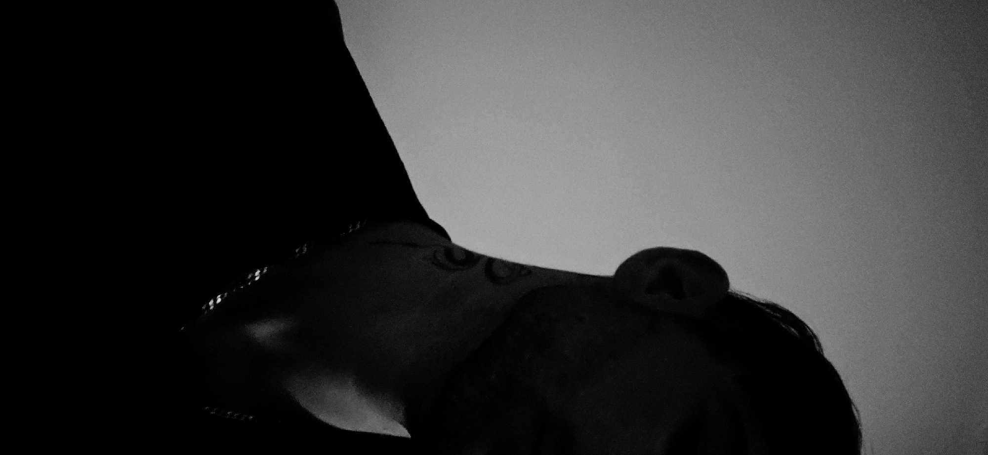 The image is a low-angle, black and white photograph. The subject is a person, possibly wearing a dark garment or hood that obscures the left side of the frame. A portion of the person's face, including an ear, cheek, and what appears to be a tattoo on the neck, is visible. The background is a light grey or white gradient, suggesting an overcast sky or a blank backdrop. The photo is underexposed and lacks detail in darker areas. There is no evidence to determine a specific location, including Plovdiv, Bulgaria.