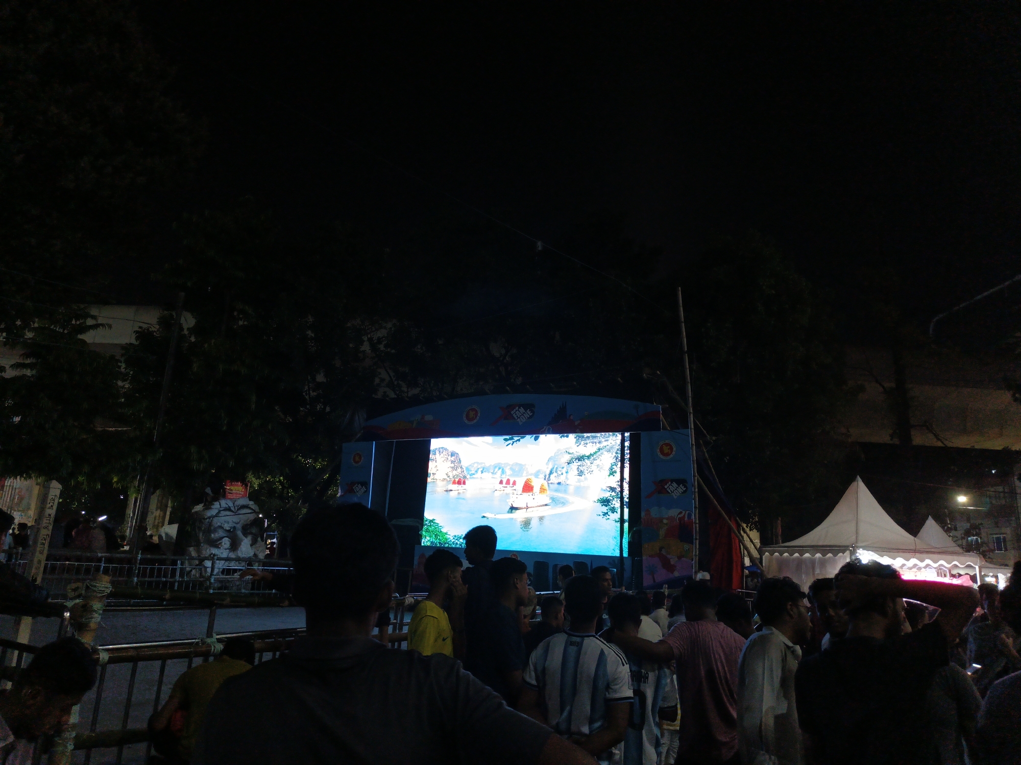 A crowd of people are gathered outdoors at night, watching a large LED screen that displays a scenic view of boats sailing on a body of water surrounded by karst formations. The screen is part of a larger structure with colorful banners, one of which says "FAN ZONE" and another featuring what appears to be a country's flag or emblem. The scene is lit by the glow of the screen and some ambient lighting, casting long shadows and creating a somewhat dark atmosphere. The people in the foreground are silhouetted against the screen, and some are wearing jerseys, suggesting a possible sporting event or fan gathering. The overall mood appears to be one of communal engagement, with individuals facing the screen and interacting with each other. Behind the crowd, tents are visible, and trees with foliage are silhouetted against the night sky. The time of day is clearly night. The weather appears to be clear, as there are no signs of rain or extreme conditions.