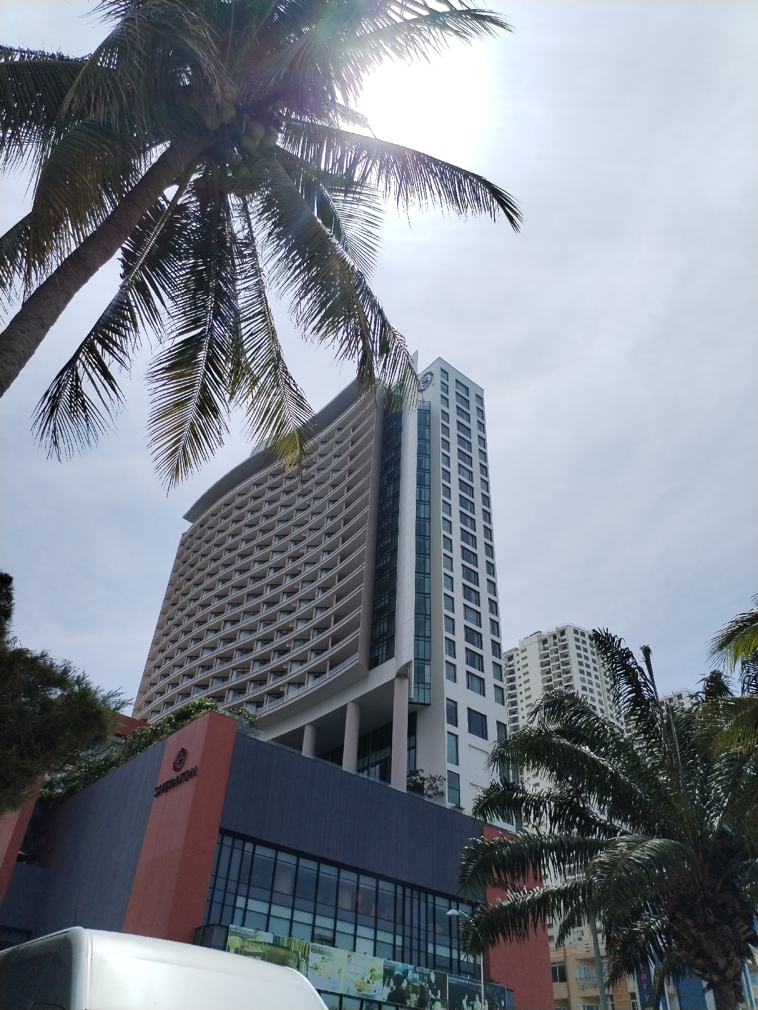The image depicts a wide-angle view of a modern hotel building in Nha Trang, Vietnam, likely located in a coastal area given the presence of palm trees. The hotel appears to be a multi-story structure with a distinctive curved facade on the upper levels, characterized by numerous balconies and windows. A more slender, white high-rise building stands to the right of the primary hotel, with another building further in the background. The foreground is partially obscured by lush green palm trees, with their fronds casting shadows and framing the architecture. The sky is overcast, with a bright glare suggesting the sun is behind the clouds, creating lens flare effects in the upper portion of the image. The lower section of the main hotel features a dark facade with the "SHERATON" logo prominently displayed on an orange section. A white vehicle is partially visible in the bottom left corner, suggesting it's parked or passing by. The overall impression is of a contemporary urban landscape blended with tropical vegetation.