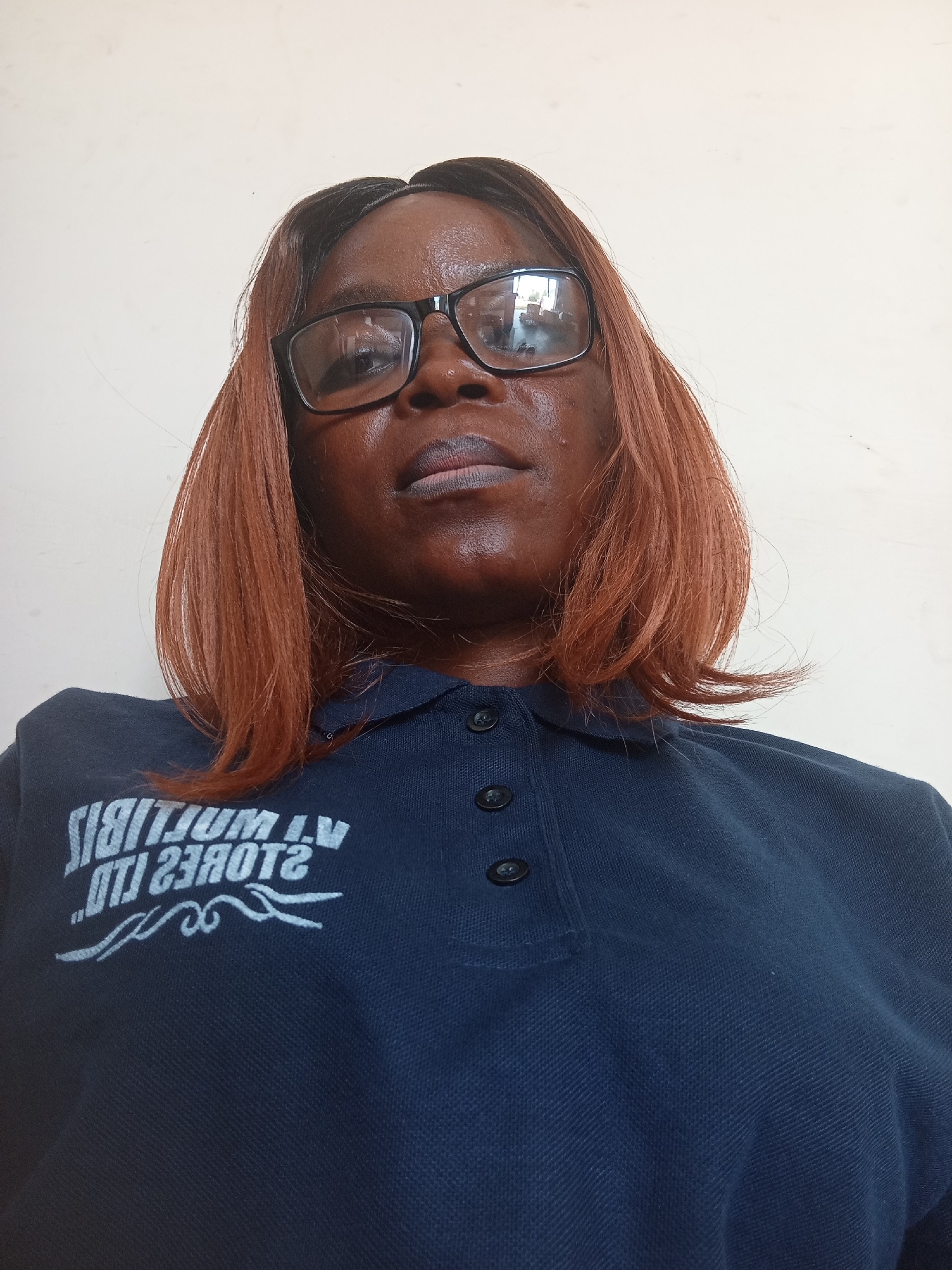 The media file shows a close-up, low-angle selfie of a Black woman wearing glasses and a dark blue polo shirt. The woman has medium-length, reddish-brown hair and is looking directly at the camera. Her expression is neutral, and the lighting suggests an indoor setting with overhead light.
The polo shirt has a white logo on the left side, which appears to be text and a decorative flourish. The visible text reads, "ATI WORTIJUM L.V. OTI 2380TZ". The shirt has three dark buttons at the neck.
The background is a plain, light-colored wall, likely indoors, given the lighting and absence of any outdoor elements. The low-angle perspective makes the viewer look up at the woman. The overall scene is simple and focuses on the subject. There are no explicit indications of the time of day, weather, or a specific event taking place. The location context provided is Mayo Dasa, Nigeria.