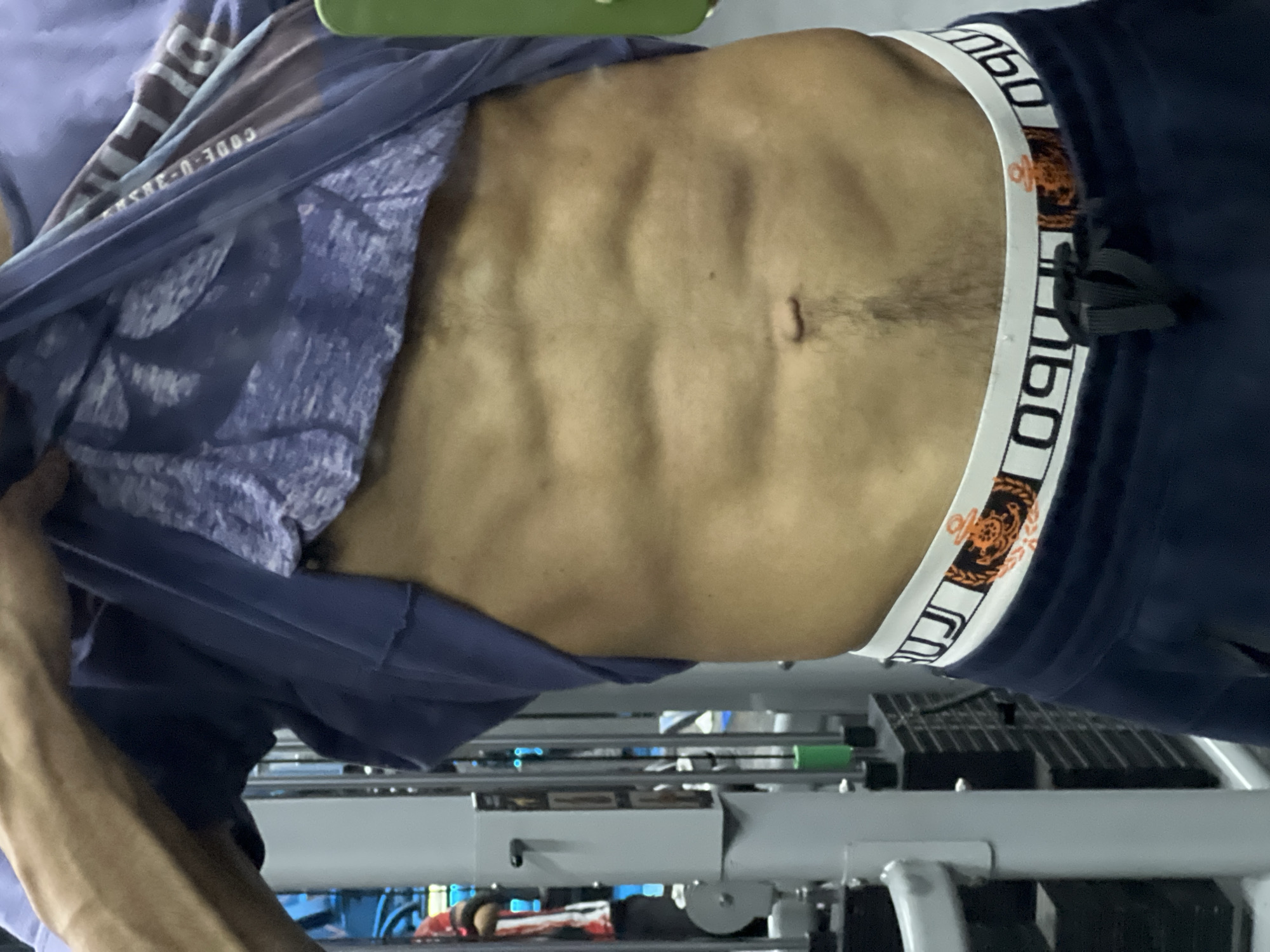A man is indoors, partially revealing his abdomen which shows muscle definition and a navel. He is wearing blue clothing including sweatpants with a white waistband featuring bold black lettering and decorative orange elements, and a purple t-shirt with text that reads "CODE U-385" and white patterns. The man is likely at a gym because exercise equipment is visible in the background. The scene appears to be in M'Diq, Morocco.