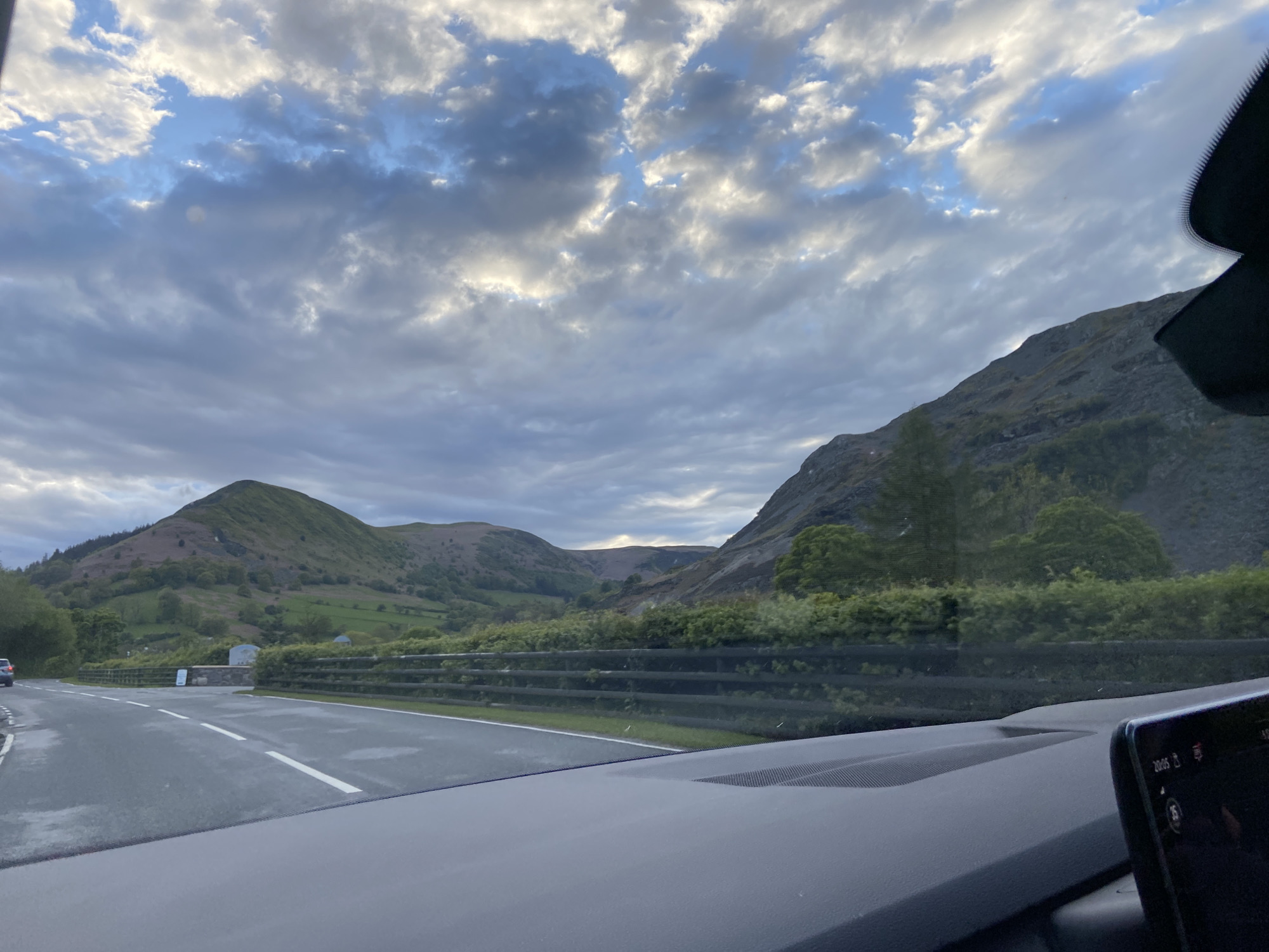 The image shows a view from inside a car, driving on a road in a mountainous region, likely in or near Llangynog, United Kingdom. The road is wet, with visible lane markings. Guardrails line the side of the road, separating it from the surrounding greenery and mountain slopes. Two mountains can be seen in the background, one with a rounded top covered in green vegetation, and the other steeper and rockier. The sky is cloudy. A portion of the car's dashboard is visible in the foreground, including a digital display showing the time "20:05." Another vehicle is driving on the opposite side of the road, in the distance.