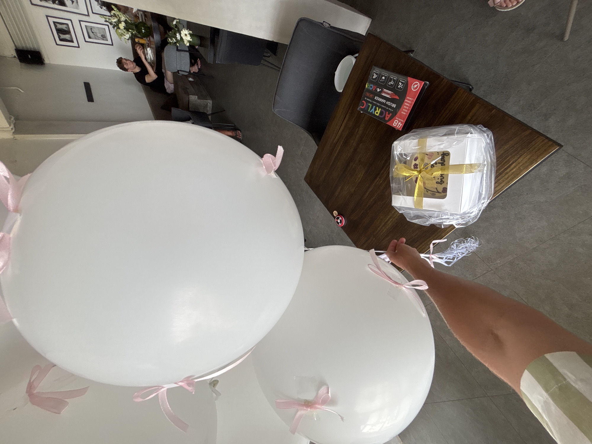 The image shows a room with white balloons and a brown wooden table. On the table, there's a box with a yellow ribbon and the word "birthday" visible, and a box of acrylic markers. A person is holding one of the balloons. The room appears to be indoors, with a view to an outside area where a few people are seated. There is no direct indication of being in Kuta Selatan, Indonesia.