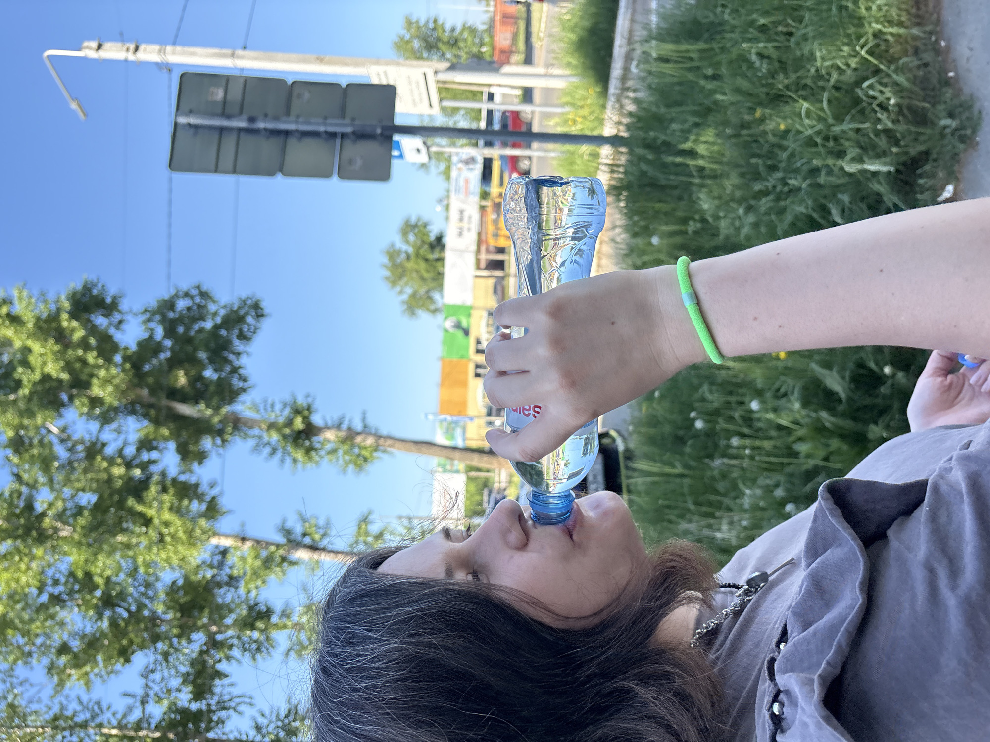 A person is drinking from a plastic bottle of water outdoors, on a sunny day in what appears to be a city setting.
The individual is holding a "Saint Springs" branded water bottle to their lips, tilting the bottle upward to drink. They are wearing a gray shirt, a silver necklace, and a lime green bracelet on their left wrist.
In the background are trees, a traffic signal, various buildings and signage, and greenery suggesting a park or roadside grass area. The location is identified as being in the city of Chaikovsky, Russia.