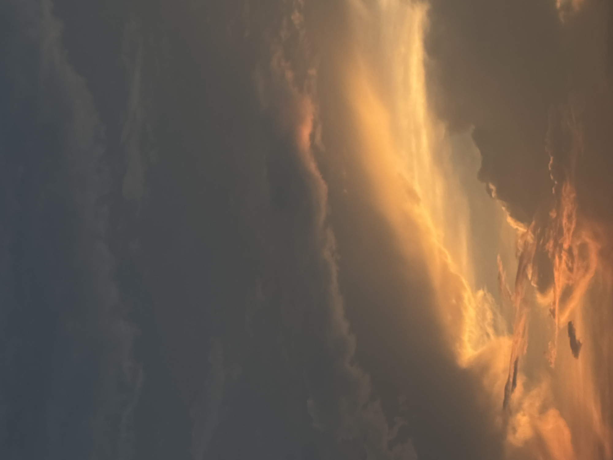 The image depicts a sky at sunset in Puebla, Mexico. Dominating the scene are dramatic cloud formations, with the left side displaying dark, dense cumulonimbus clouds. To the right, sunlight breaks through, creating a striking contrast. The sunlight is warm and golden, illuminating the edges of the clouds and casting an ethereal glow. The clouds are textured and varied, indicating a dynamic atmospheric condition. There is no evidence of people or human-made objects in the image. The primary subject is the sky itself, captured during a moment of intense light and shadow play.