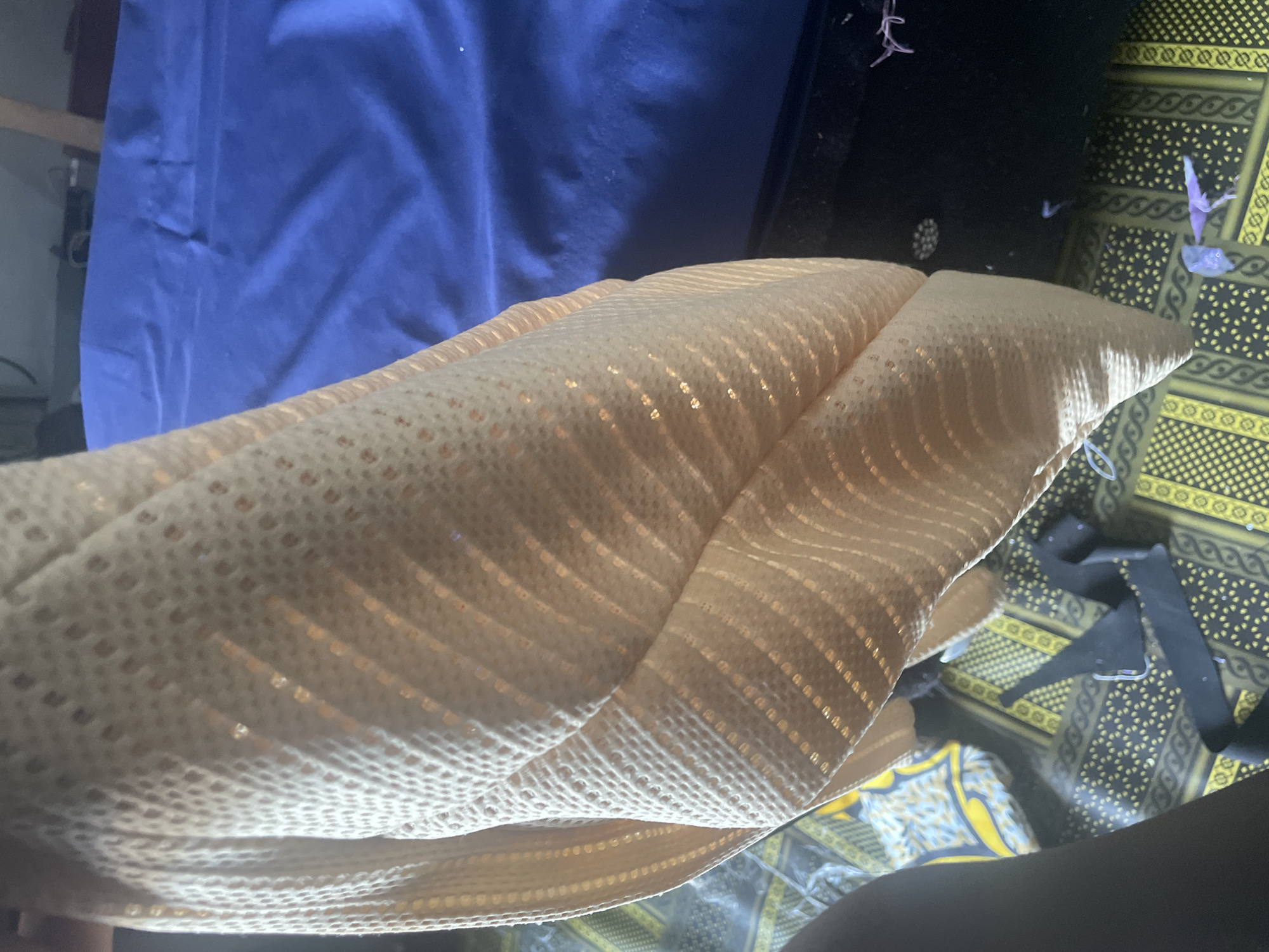 The image depicts a close-up view of a pale peach-colored fabric with a textured, perforated pattern and subtle iridescent stripes, possibly sequins or metallic threads. The fabric is draped, creating folds and shadows that highlight its texture. In the background, a portion of a bright blue surface, likely a bedsheet or blanket, is visible, contrasting with the fabric in the foreground. The floor beneath the fabric is covered with a patterned rug or textile featuring yellow and dark green geometric designs. Some black fabric scraps and small, light-colored objects, possibly craft supplies like beads or glitter, are scattered on the floor. The lighting suggests an indoor environment, possibly a bedroom or a crafting space. Without additional context or visible landmarks, the specific location within Jalingo, Nigeria, cannot be determined.