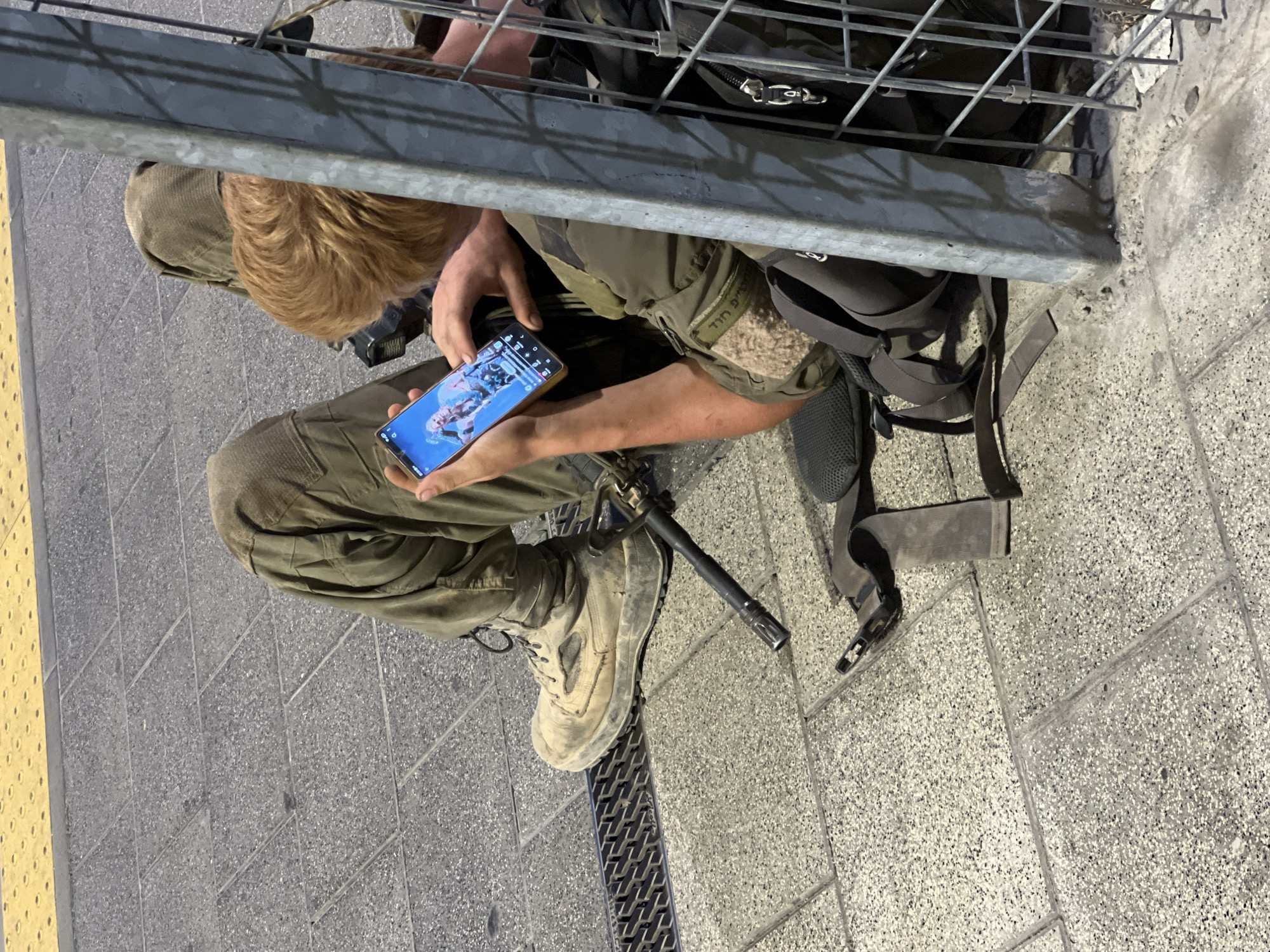 A person with reddish-blonde hair wearing military-style clothing and boots is sitting on the ground in what appears to be an urban environment. They are looking at and interacting with a smartphone, which displays an image of a person. A rifle lies next to their left leg, and a backpack is positioned behind them. The ground is paved with grey interlocking tiles and has a yellow tactile paving strip along the top edge, indicative of a sidewalk or platform. A metal grate is visible on the left side of the frame, and a metal pole and fence are to the right, suggesting a public space possibly near a transit area. The person's clothing has patches with Hebrew writing, and the overall scene is consistent with a location in Haifa, Israel.