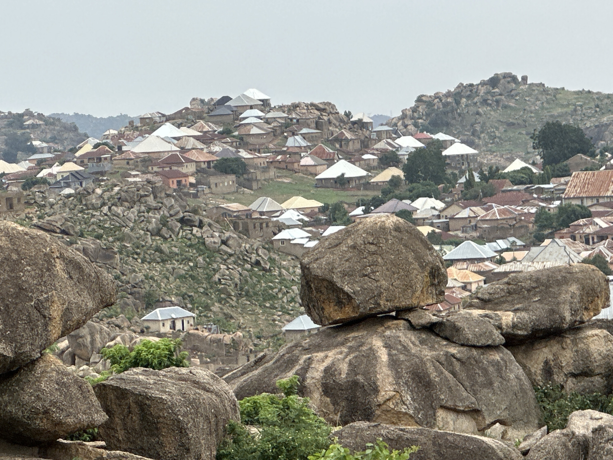 The image depicts a densely populated hillside in Jos, Nigeria. The foreground consists of a cluster of large, weathered, gray-brown boulders, some topped with greenery. Behind these rocks lies a community of closely-packed buildings, most with corrugated metal roofs in shades of silver, white, and rust. The buildings vary in size and appearance, suggesting a mix of residential and commercial structures. The structures are built on and around rocky terrain, with a mix of bare rock, scrub vegetation, and trees visible. In the background, a larger, rockier hill rises, also densely populated with buildings. The sky is overcast and hazy, indicating a diffuse light and limiting visibility of distant features. No people are discernible in the photograph.