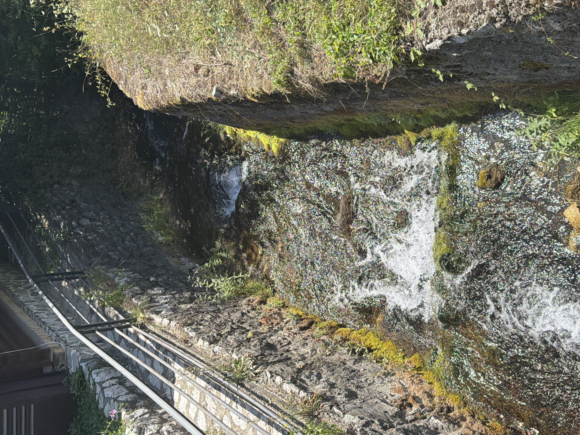 The image depicts a narrow, stone-lined channel of flowing water. The channel walls are constructed of rough, dark stones, with patches of green moss and vibrant yellow-green algae growing along their edges. Vegetation, including grass and small plants, grows along the top of the right-hand wall, some of it overhanging the water. On the left side, a metal railing runs along a stone pathway that borders the channel. The water itself is clear and appears to be flowing swiftly, with white frothy areas indicating turbulence. Sunlight reflects off the surface of the water, creating a sparkling effect. In the background, the channel narrows and disappears into shadow, hinting at a continuation of the waterway. The overall impression is one of a natural watercourse that has been channeled and bordered by man-made structures, likely within an outdoor, possibly historical or scenic location.