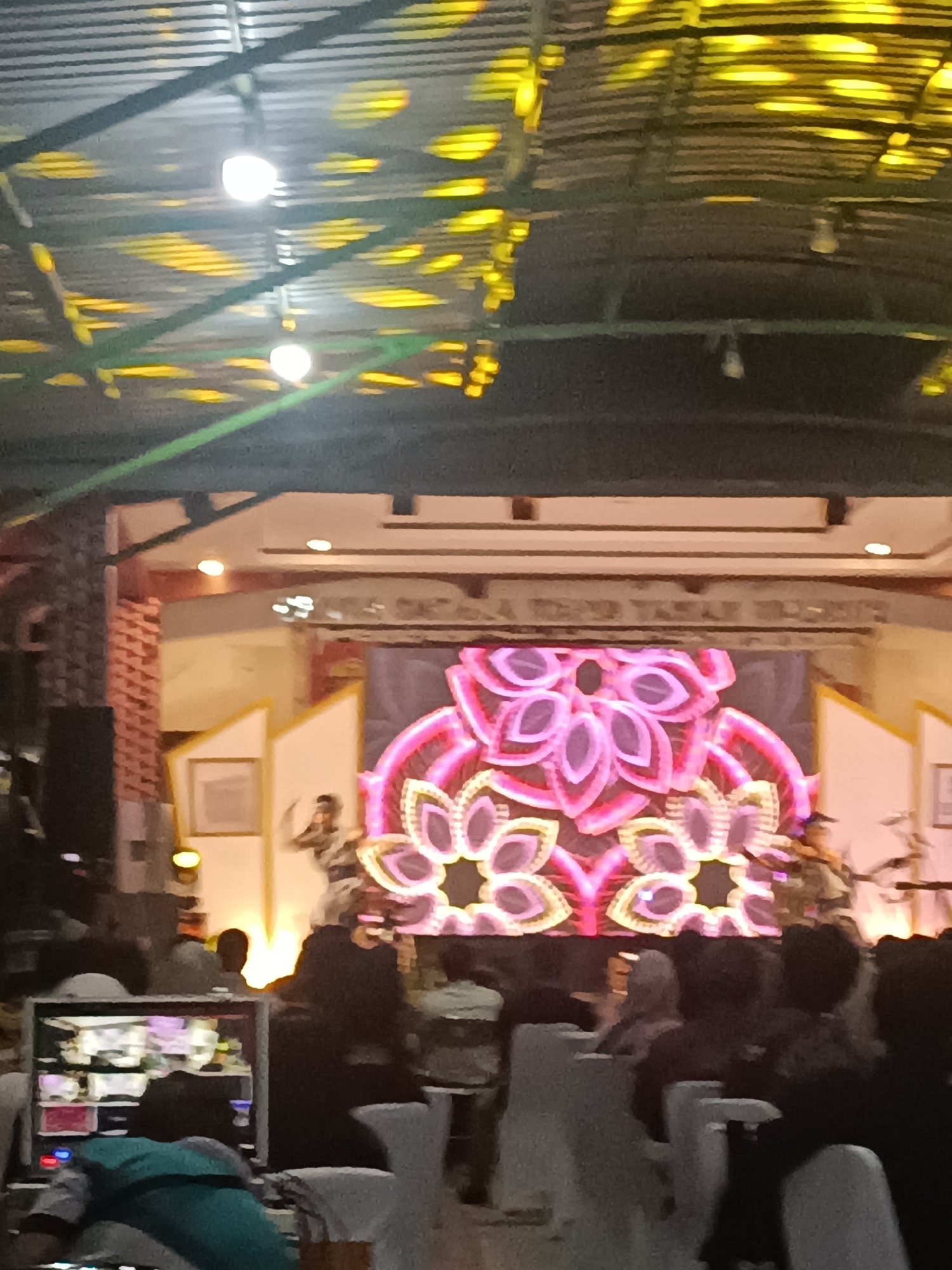 The image depicts a performance taking place indoors, likely on a stage with a large LED screen displaying a vibrant, floral-like pattern in shades of pink, purple, and yellow. Several individuals, presumably performers, are visible on the stage, some in motion. In the foreground, an audience is seated, facing the stage. To the left, a monitor displaying various visual inputs is positioned, suggesting a production or broadcasting setup. The ceiling features a corrugated metal roof with visible support beams and lighting fixtures casting yellow patterns on the surface. The overall environment appears to be a venue or hall, potentially for cultural events or performances. The location is identified as Condongcatur, Indonesia.