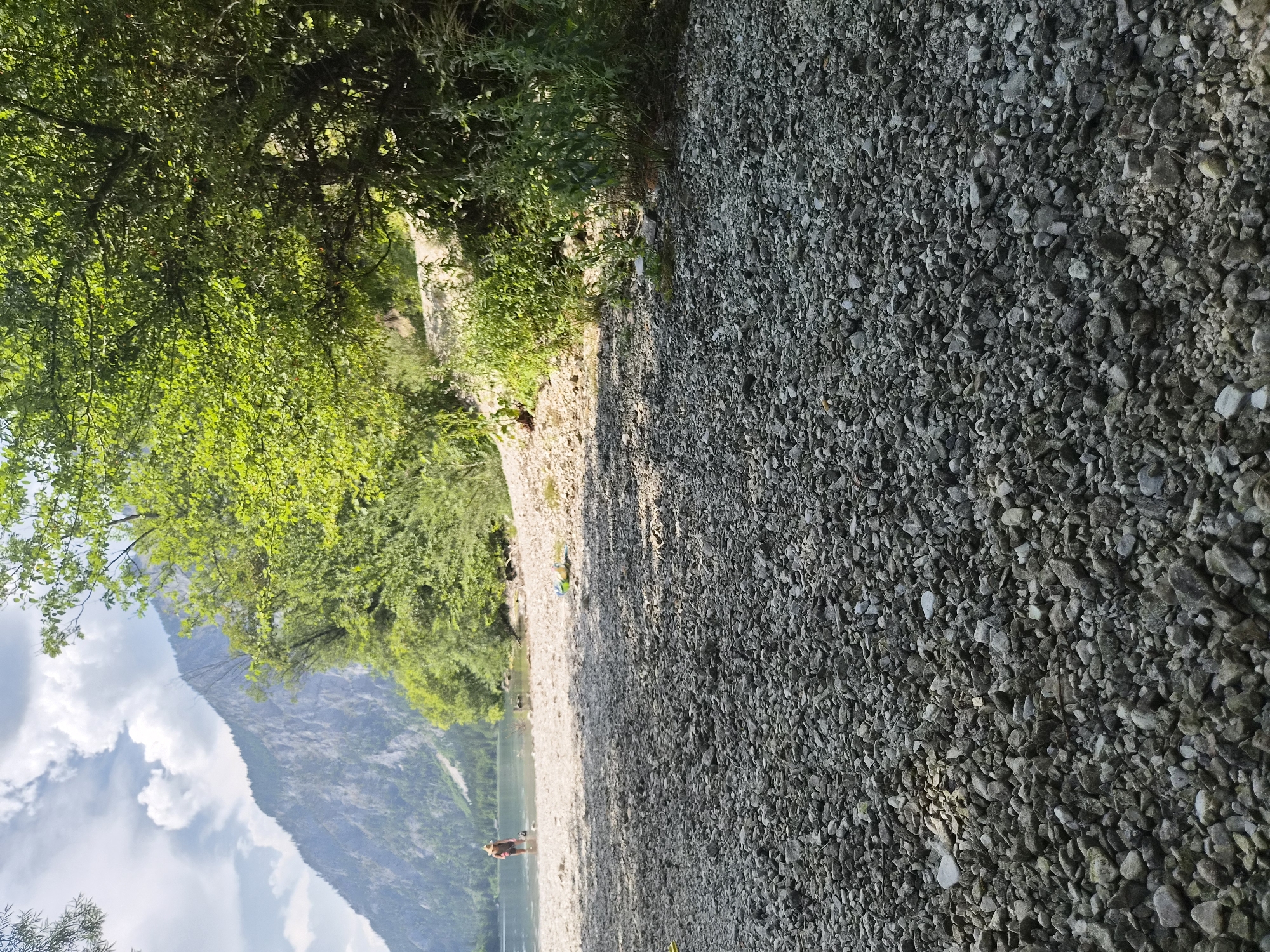 The image depicts a rocky shoreline of a lake in Eisenerz, Austria. In the foreground, a dense scattering of grey and white pebbles covers the ground. Beyond the pebbles, a person wearing a hat stands in the shallow water. Trees and lush green vegetation line the background, with a steep, rocky mountain rising in the distance under a partly cloudy sky. The scene appears to be a natural recreational area.