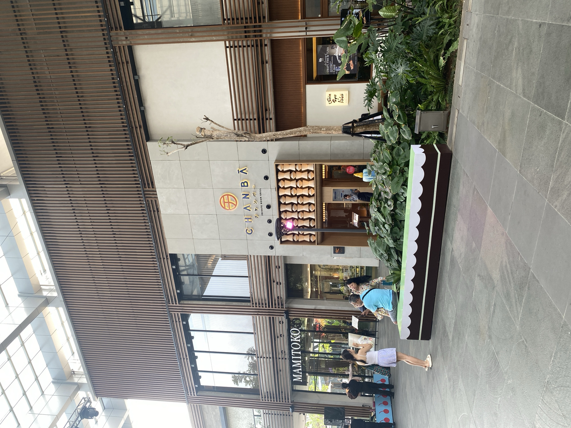 The image shows a street-level view of a modern building with a light-colored stone facade and wooden slat accents. The building houses several establishments, including "MAMITOKO" and "CHANBA PRIVATE ROOM GRILL". A woman in a white tank top and striped shorts stands on the left, holding a phone and looking at something off-camera. A man in a blue t-shirt and shorts is seated on a bench-like structure resembling a cake, with a woman in a floral dress next to him. The bench is decorated with green and brown stripes and a white scalloped edge. Lush green plants are arranged in front of the establishments. The ground is paved with grey stone tiles. The lighting suggests it is daytime. The image was taken in North Jakarta, Indonesia, likely at a commercial or entertainment district.