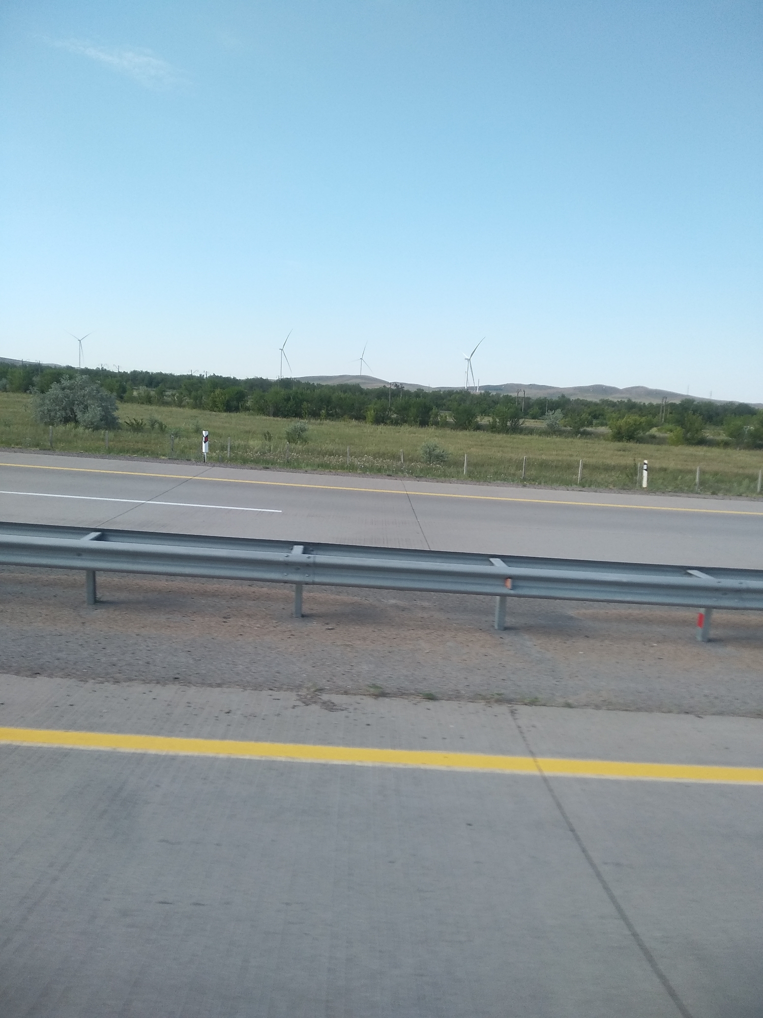 This is a photograph taken from a vehicle, likely on a highway, looking towards a grassy field with a line of trees in the distance. Several wind turbines are visible on a rolling, hilly horizon. A guardrail runs along the side of the road in the foreground. The sky is a clear, light blue. The scene is set in the vicinity of Osakarovka, Kazakhstan, suggested by the presence of wind turbines in a landscape typical of the region. There are no people or animals visible in the frame. The overall impression is one of a rural, open landscape under a clear sky.
