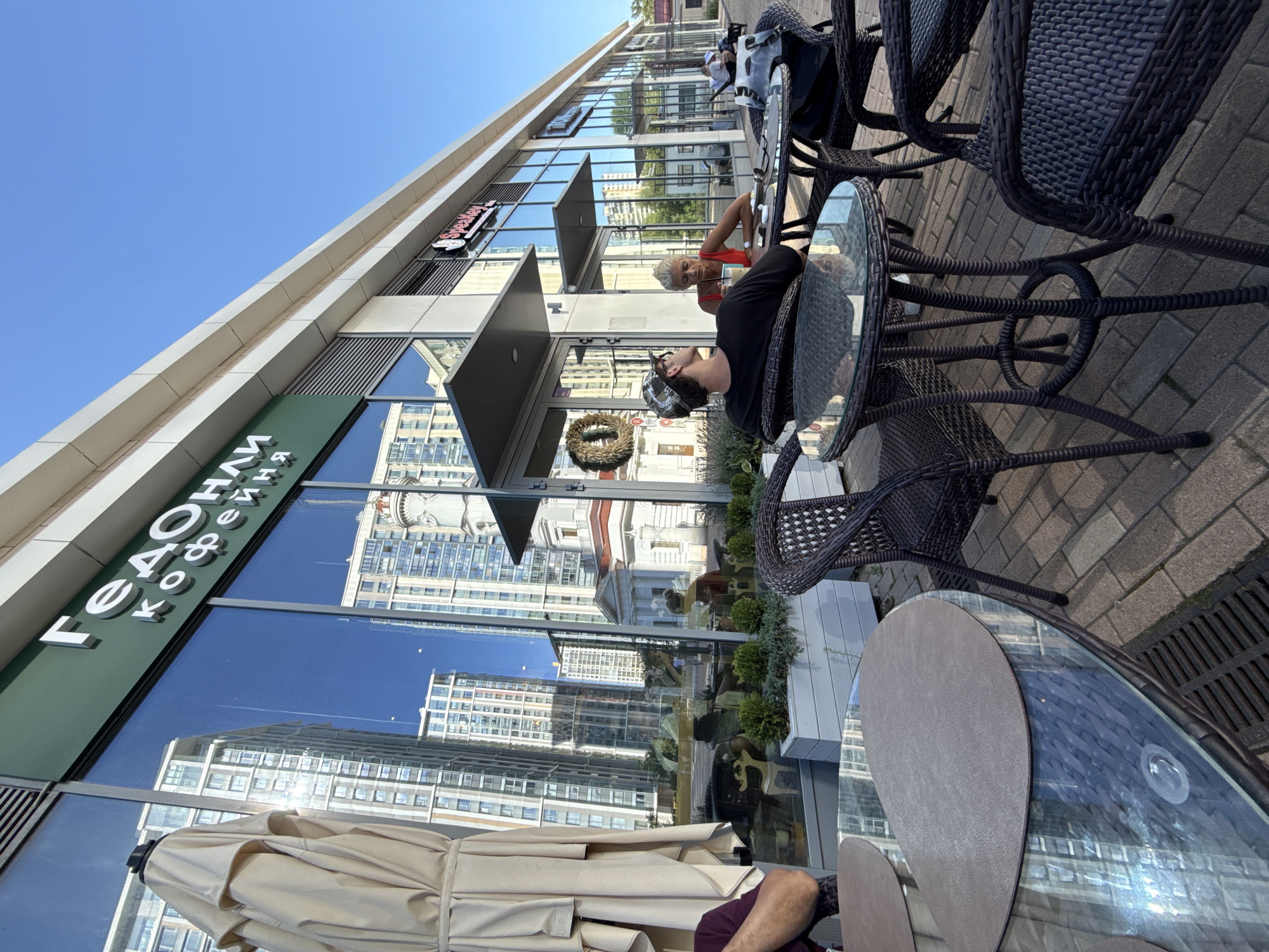 The image captures an outdoor seating area of a cafe, likely located in Moscow, Russia, given the Cyrillic signage that reads "Гедонист Кофейня" (Gedonist Coffee House). The cafe is situated along a modern street with a glass facade reflecting tall, contemporary apartment buildings and a white classical-style building with a clock tower.
In the foreground, two people are seated at a round, glass-topped table with wicker chairs. A young man wearing a baseball cap faces away from the camera, looking towards a woman seated opposite him. The woman, with blonde hair, is wearing a red top and appears to be holding a drink. Another table with chairs is visible to the left, with a partially visible person seated. To the right, more tables and chairs are arranged, with other patrons in the distance.
The setting suggests a pleasant, sunny day, with clear blue skies overhead. Planters with greenery are placed along the facade of the building, adding to the ambiance of the outdoor cafe. The overall scene depicts a casual, urban environment where people are enjoying refreshments in a relaxed atmosphere.