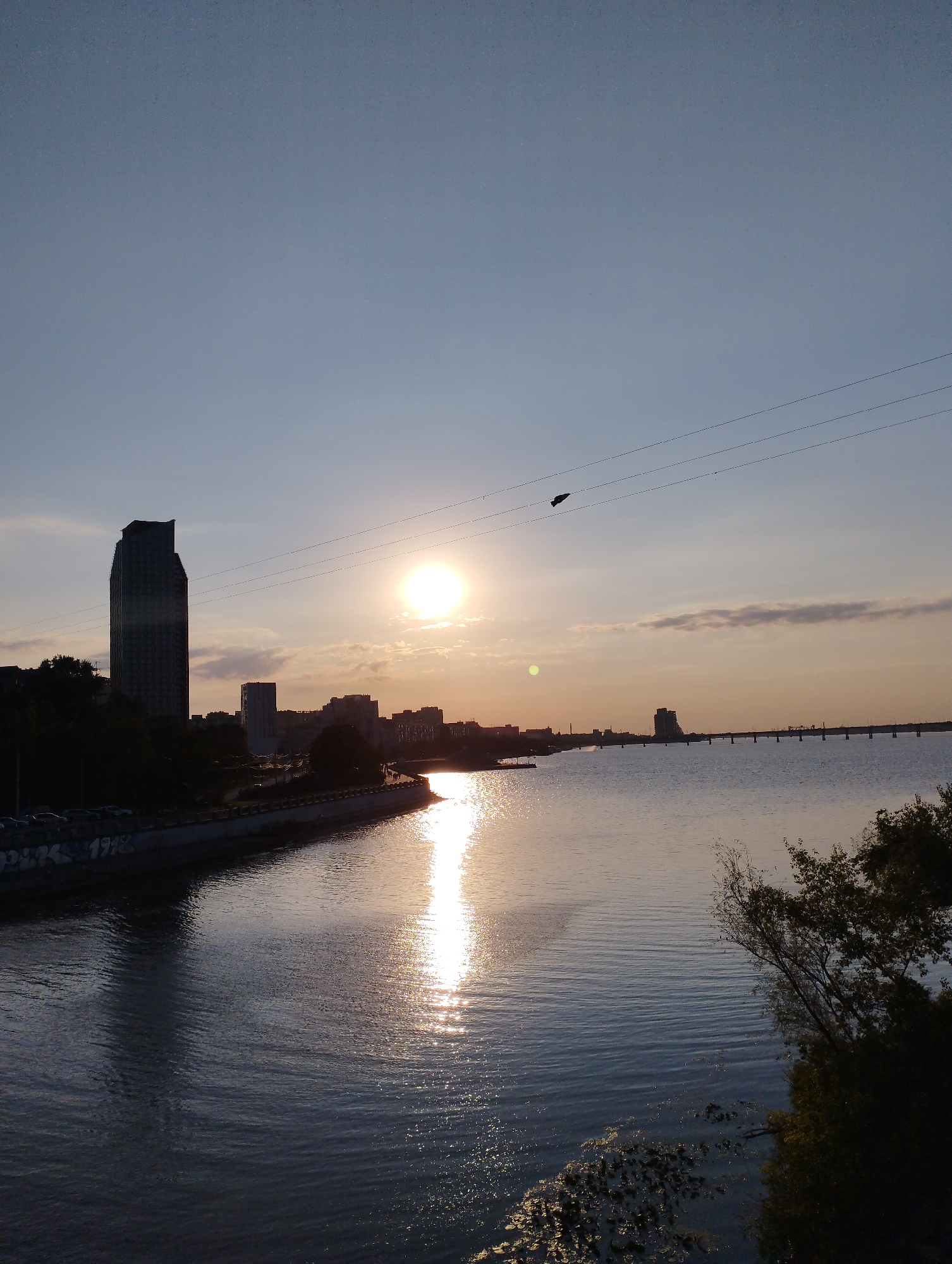 A cityscape at sunset is depicted from an elevated perspective, overlooking a wide body of water. The sun, a bright orb low in the sky, casts a shimmering reflection across the water's surface. The shoreline features a promenade lined with trees, a tall, modern skyscraper, and other buildings forming a city skyline. A bridge extends across the water in the distance. Power lines are visible strung across the sky. A single dark object, possibly a bird or an object on the power lines, is visible in the upper part of the frame. The water is calm with gentle ripples. The overall scene is tranquil, bathed in the warm, fading light of sunset.