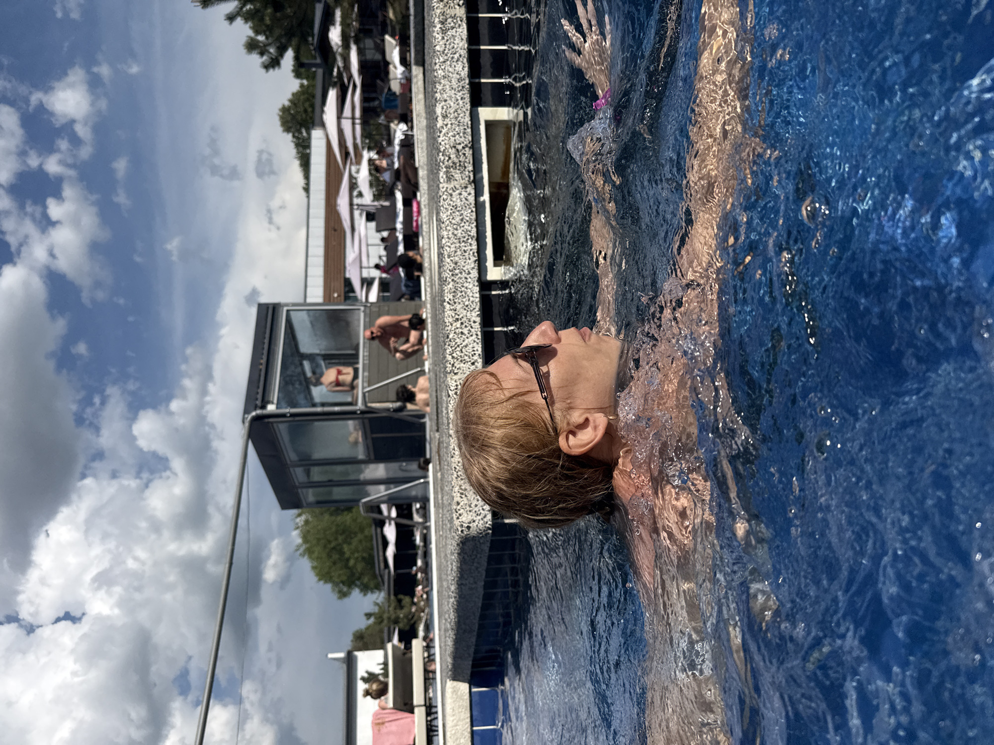 A person is submerged in a blue swimming pool, up to their neck, on a sunny day. The individual is wearing sunglasses and has short, light-colored hair. The pool's edge is visible, with a gray and white stone border and blue tiles. In the background, there's a multi-story building with glass walls, lounge chairs, umbrellas, and people sunbathing. The sky is blue with scattered white clouds. No information is available regarding the location in the city of Rostov-on-Don, Russia.