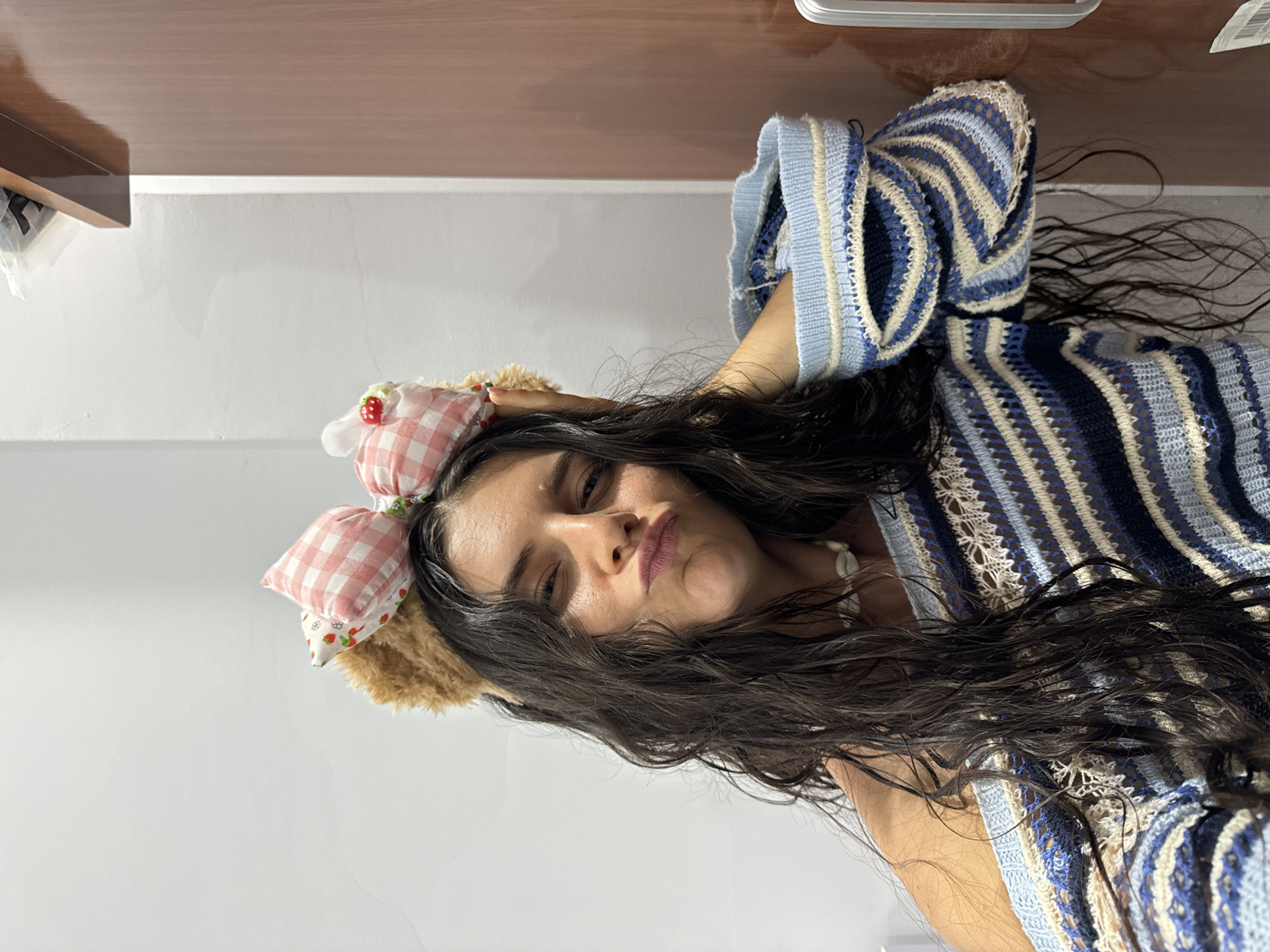The image features a young woman in a close-up, chest-up shot, presented in a portrait orientation (rotated clockwise in the original file). She has long, dark, and wavy hair that frames her face and flows over her shoulders and chest. Her expression is playful, with slightly puckered lips and one eye partially closed as she looks directly at the camera, conveying a cheeky or flirtatious mood.
She is wearing a distinctive knitted or crocheted top featuring wide horizontal stripes in shades of light blue, dark blue, and beige, with an intricate open-knit or lacy pattern integrated into the design. Adorning her head is a headband with unique embellishments; it has two prominent, plush, pink-and-white checkered bow-like elements on one side, one of which features a small red strawberry appliqué. A section of the headband closer to her ear appears to be made of a fluffy, light brown, fur-like material, possibly resembling animal ears. Around her neck, a necklace made of small, white, shell-like beads is visible.
The background is simple and uncluttered, primarily consisting of a plain, light-colored wall. Above this wall, a band of a smooth, dark wooden surface, likely a shelf or furniture, is visible. The lighting is soft and even, indicative of an indoor setting or a shaded area. No discernible text is present in the image.