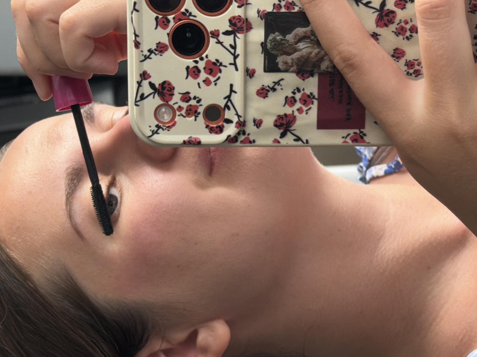 A woman is seen applying mascara to her eyelashes while holding a smartphone with a floral pattern. The smartphone is angled to capture her reflection. The background is blurred, but it appears to be an indoor setting. There are no discernible landmarks or indicators to suggest a specific location within the city of Sochi, Russia.