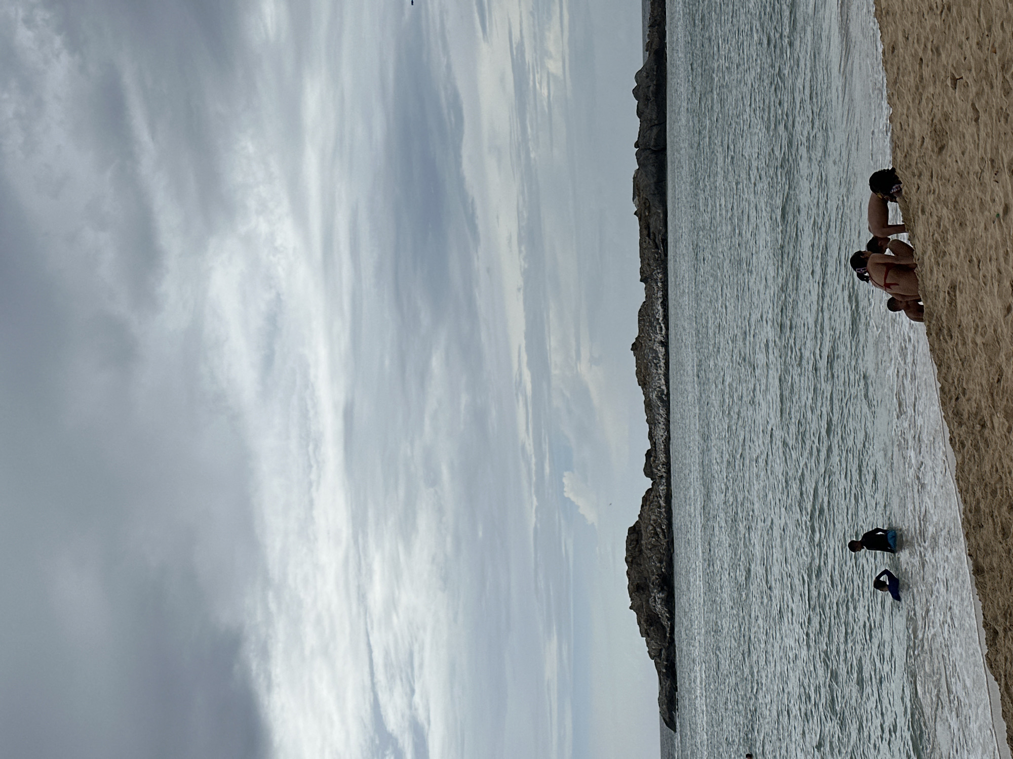 The image depicts a scene at a beach with a cloudy sky and a body of water. The perspective is from the sand looking out towards the sea and the sky. There are several people visible in the water and on the beach.
In the foreground, on the right side of the image, is a sandy beach. There are a few people sitting or crouching on the sand near the water's edge. One group appears to be interacting or observing something in the water.
In the water, there are at least two individuals on what appear to be bodyboards. One person is wearing a dark wetsuit and is further out in the water, while another person in a dark blue wetsuit is closer to the shore. The water is calm with a textured surface, suggesting it might be shallow or that there are gentle ripples.
The background shows a rocky coastline or cliff face extending into the distance. The sea stretches out towards this rocky area, and beyond that, the sky is filled with numerous clouds. The clouds are streaky and varied in density, with some areas appearing brighter than others, possibly indicating patches of sunlight trying to break through. The overall lighting suggests it might be daytime, perhaps late afternoon or a generally overcast day. The mood conveyed by the image is one of a relaxed beach outing. There is no visible text in the image.