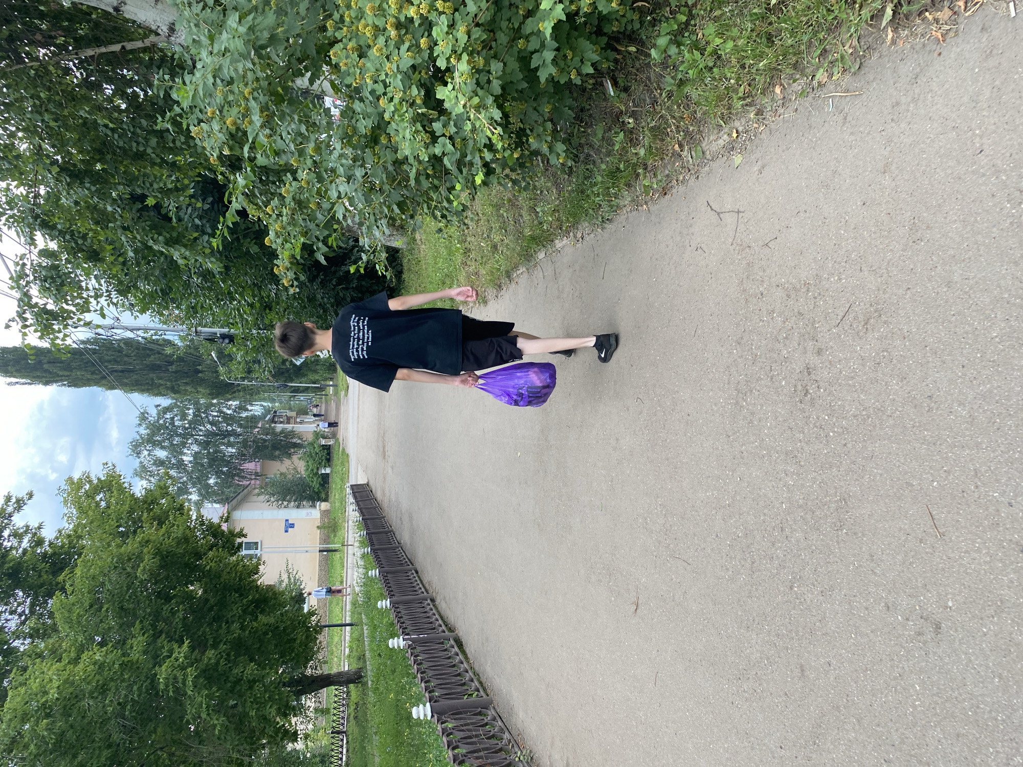 A young boy wearing a black t-shirt with white text and black shorts walks away from the camera on a paved pathway. He is carrying a crumpled purple plastic bag in his right hand. The pathway is lined on the left by a decorative metal fence and a grassy area with trees. On the right, the pathway is bordered by dense green foliage and trees. In the background, buildings and more trees are visible under a cloudy sky. The scene appears to be in a park or a residential area within the city of Ishimbay, Russia, given the architecture and vegetation.