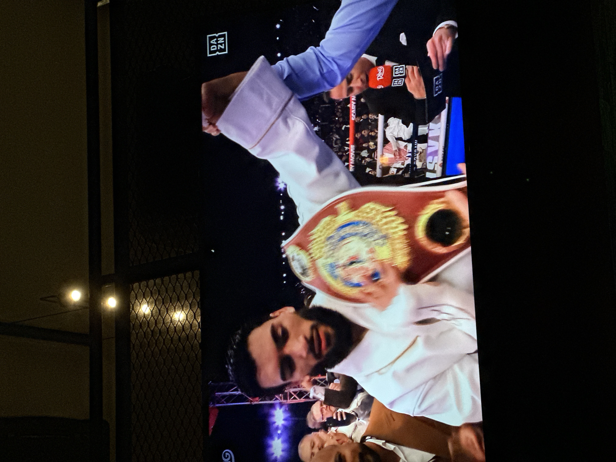 A television screen displays a boxing match aftermath. The newly crowned champion, with a beard and dressed in white, holds the championship belt aloft. A DAZN logo is visible in the upper corner of the screen. The background includes members of the champion's entourage. The image appears to be captured indoors, possibly in a residence, as evidenced by the partially visible window with a security grill and reflected light sources on the left. The photo seems to be taken in North Kuta, Indonesia.