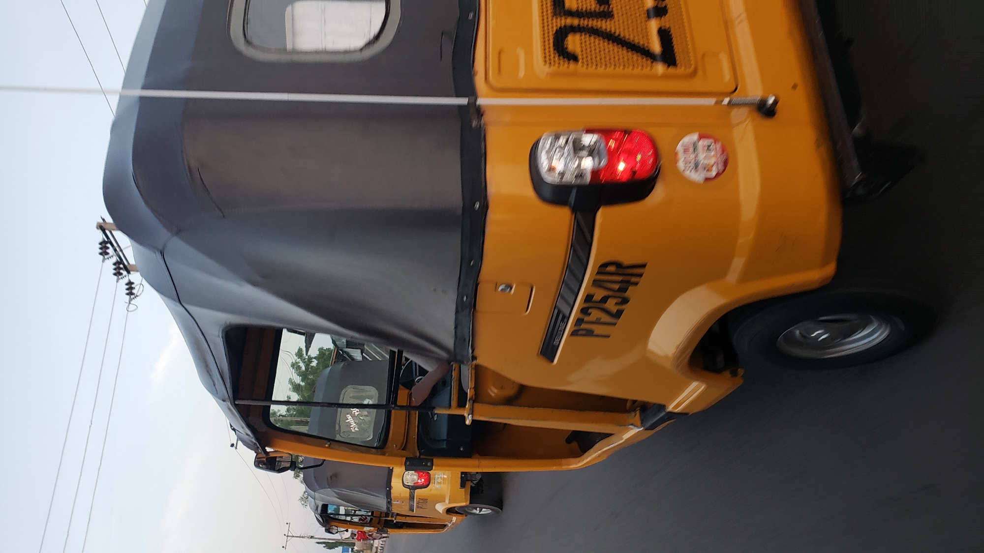 This is a close-up, low-angle outdoor photograph of three yellow auto-rickshaws (also known as Keke Napep or Marwa) driving on a paved road in Jalingo, Nigeria. The focus is on the rear and side of the closest auto-rickshaw, which displays the license plate "PT254R" and the word "DELUXE" in black lettering. A red taillight is visible on the rear of this vehicle, along with a circular sticker below it. The second auto-rickshaw, with license plate "PT1760A", is visible to the left and slightly ahead of the first. The third auto-rickshaw is further ahead and less distinct. Electrical wires are strung overhead. The sky is overcast and light. The road surface is dark asphalt. The vehicles are in motion, as suggested by the blur of the wheels.