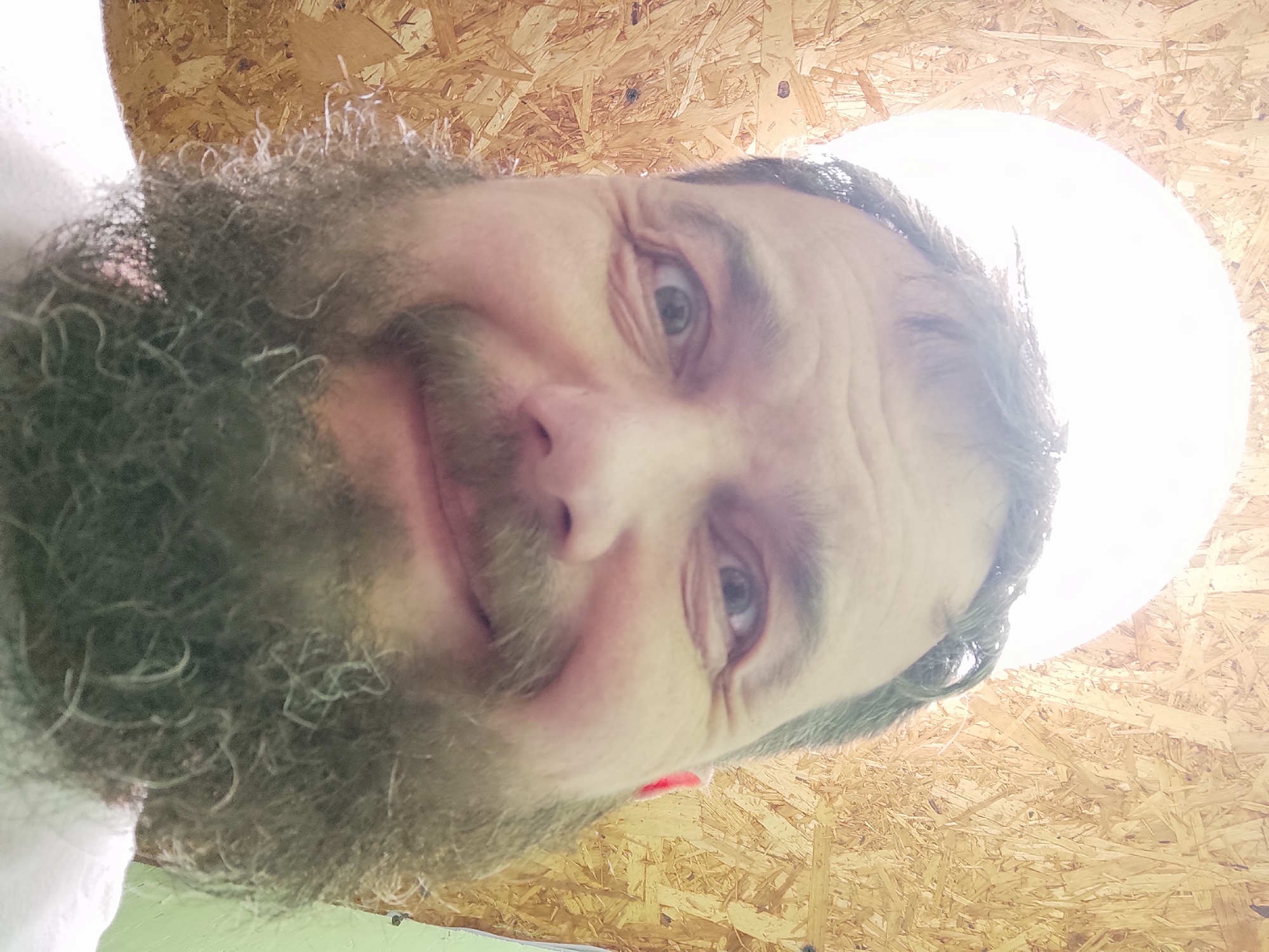 A close-up, low-angle, wide selfie of a man with a beard is taken in an indoor environment. The man is looking directly at the camera with a slight smile. The background consists of particleboard walls and a bright light source that appears to be a ceiling fixture or a circular opening. The lighting is intense, creating a blown-out effect in the upper portion of the image. The man's face is in focus, with his beard prominently displayed.