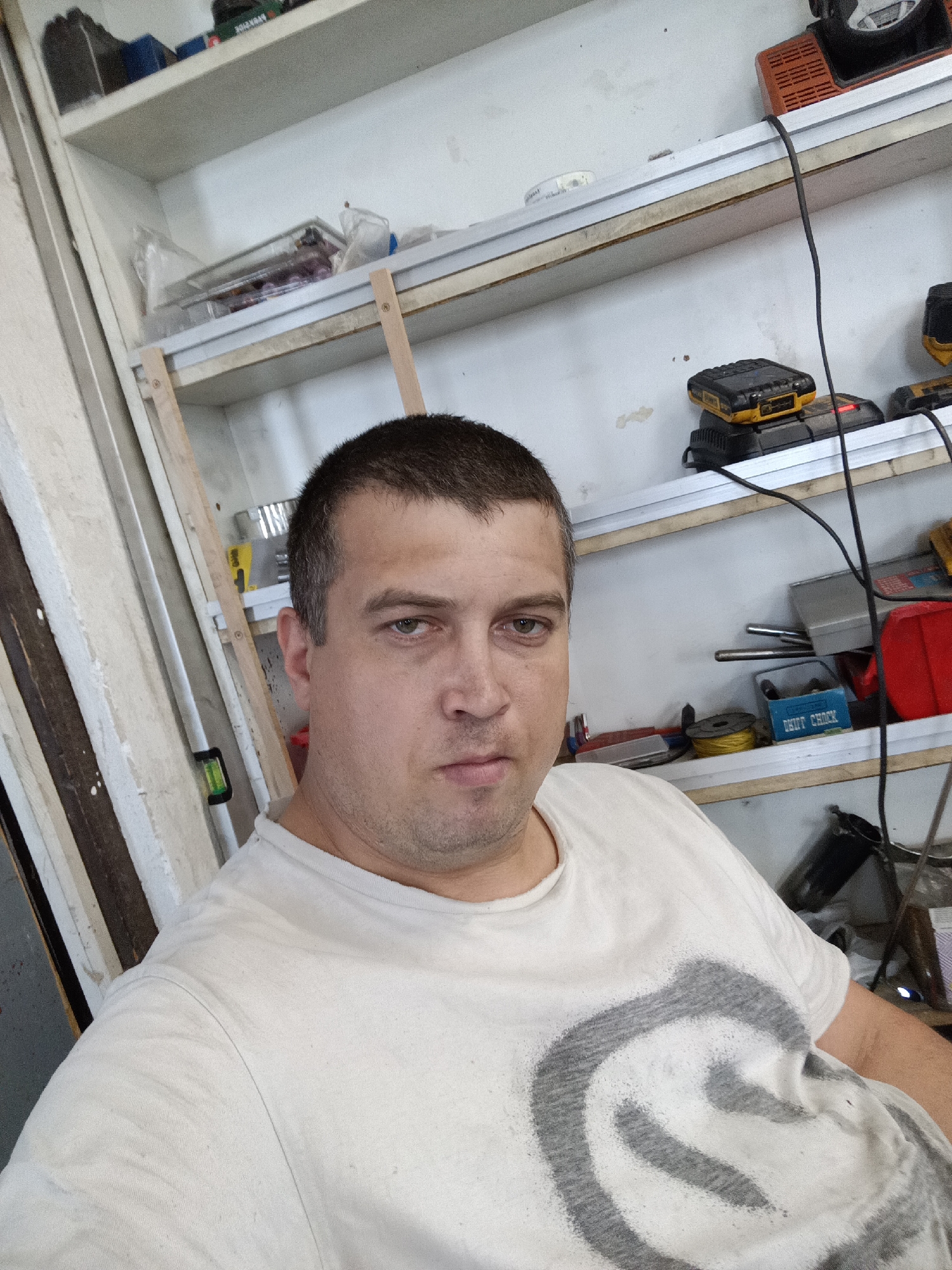 The image is a selfie taken by a man with short brown hair and fair skin, wearing a white t-shirt with a grey circular design. He has a neutral expression and is looking directly at the camera. He is indoors, likely in a workshop or garage, given the presence of shelves filled with various tools and equipment in the background. These items include what appear to be power tools, batteries, toolboxes, and other miscellaneous items. The lighting is adequate, illuminating the man and the immediate surroundings. The overall setting suggests a functional, possibly cluttered, workspace. The image does not contain any specific details that would allow for localization to Dalovy, Czechia.