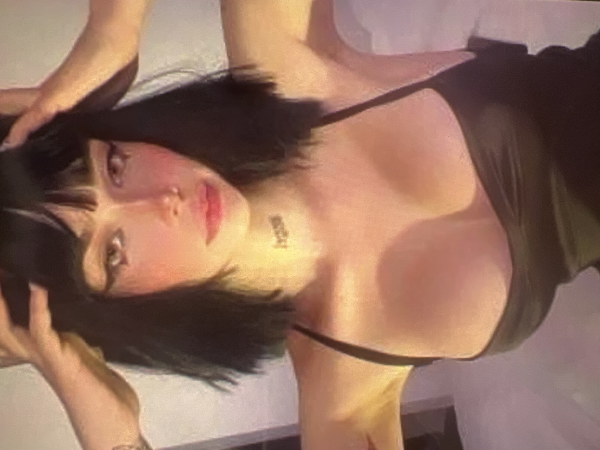 The image shows a close-up of a young woman with dark hair and bangs. She is wearing a black tank top and has her hands on her head. She has dramatic eye makeup, including winged eyeliner and false eyelashes, and her lips are painted a bright pink. There appears to be a tattoo on her neck. The background is out of focus and appears to be indoors, with soft lighting. There is no visible indication of the city of Minsk, Belarus in the image.