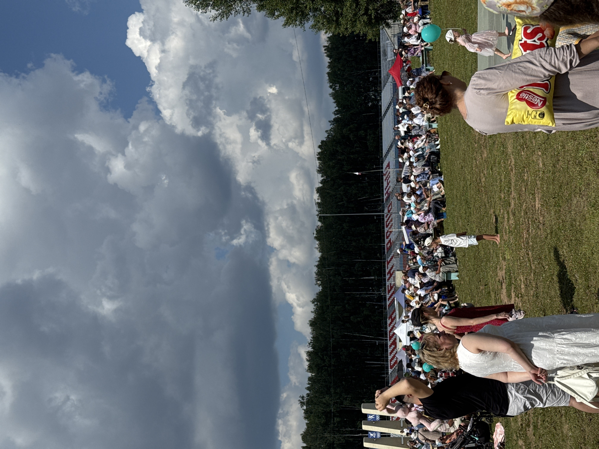 The image depicts an outdoor gathering with a large crowd seated in bleachers in the background, facing a grassy field. In the foreground, several people are visible. To the left, a man in a black t-shirt and grey shorts stands with a light pink stuffed animal held above his head. Next to him, a woman in a white dress carries a white bag. Further to the right, a woman in a red dress is partially visible. In the mid-ground, a young girl in a pink dress walks across the field holding a green balloon. Another woman in a grey dress stands to the right, with two yellow packages, one labeled "Nestle", attached to her back. The environment is a sunny day with dramatic clouds in the sky, and a line of trees forms a backdrop behind the bleachers. There are tents and flags visible in the background, suggesting an event or festival.