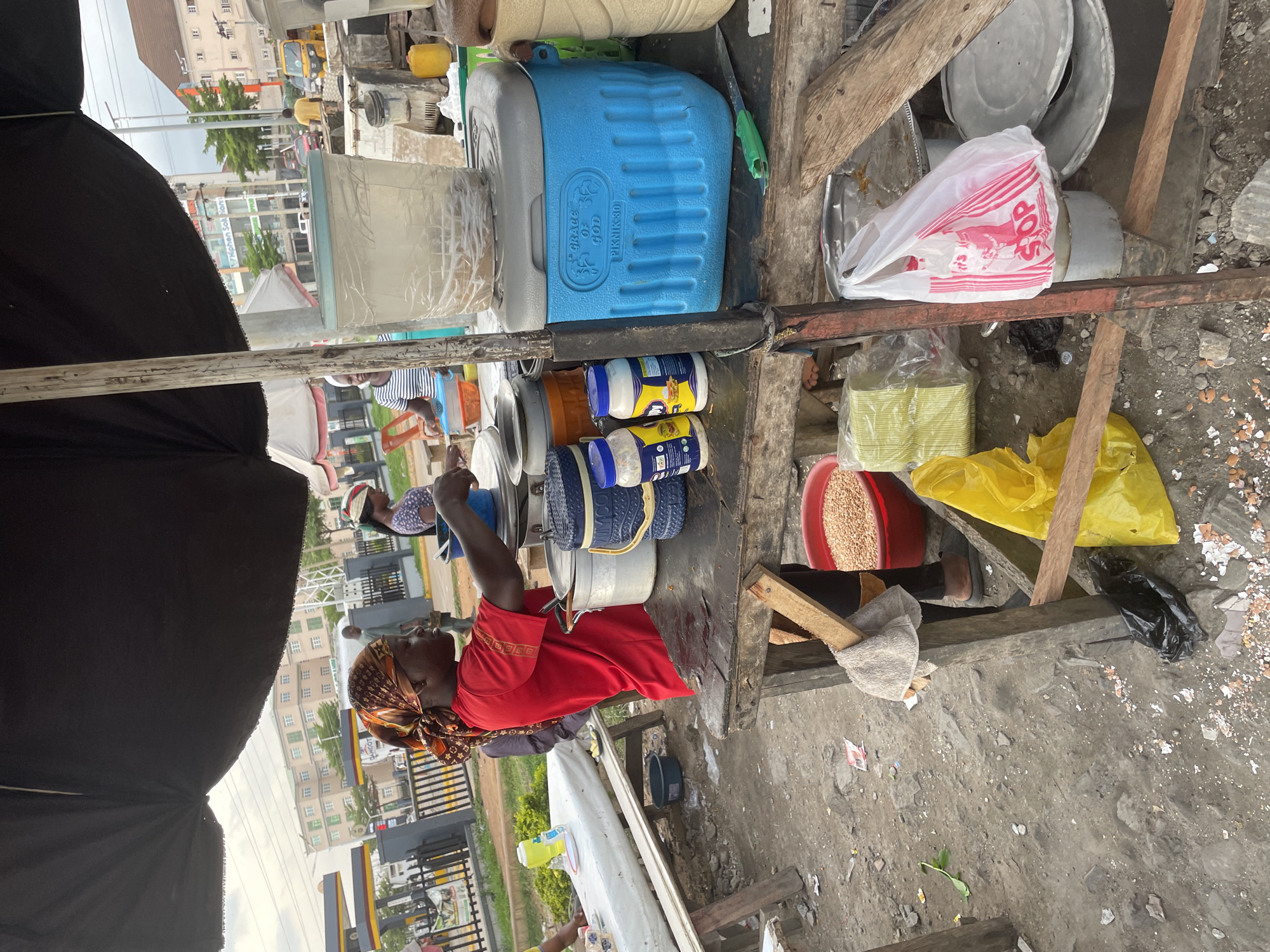 In Abuja, Nigeria, a street food vendor is preparing a meal at an outdoor stall. A woman in a red shirt and headscarf is actively serving food from pots on a wooden table. To her right, a blue cooler with the words "GRACE OF GOD" and "PIKNIK-30" is visible, topped with a clear plastic container. Other items on the table include various containers, jars of condiments, and a green knife. In the background, a road with a yellow tuk-tuk and other vehicles suggests a busy urban setting. The ground is littered with debris, including eggshells and trash. The overall scene depicts a typical day at a bustling street market.