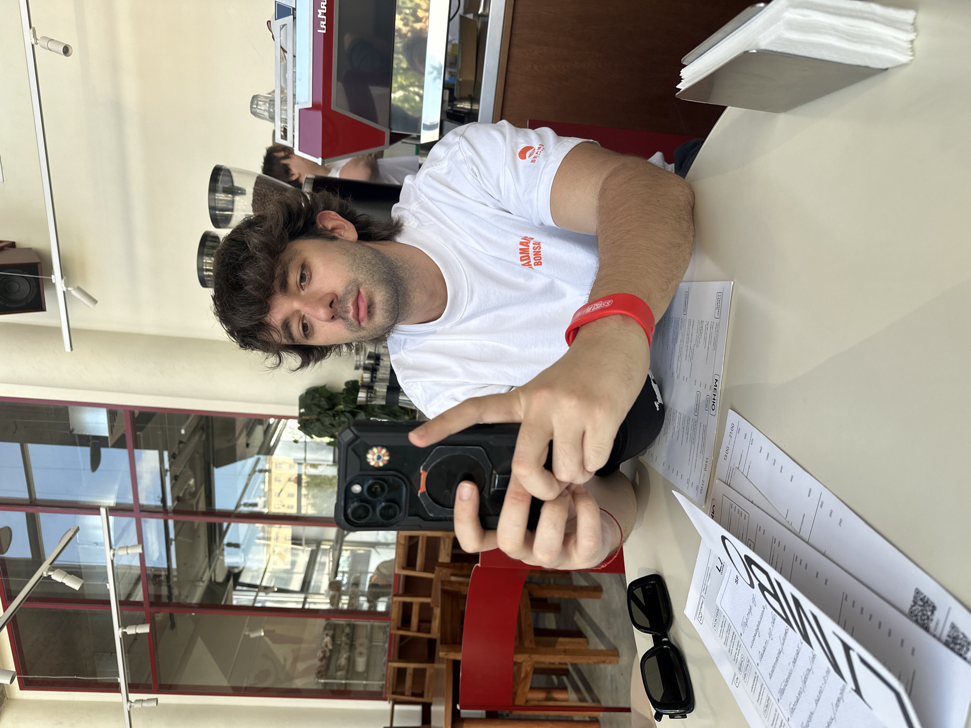 This is a vertical, medium shot of a young man in a white t-shirt and a red wristband, holding up a smartphone to take a selfie in what appears to be a cafe or restaurant. He has dark, curly hair and a serious expression. The cafe has a modern, industrial aesthetic with exposed pipes, metal fixtures, and large windows. In the background, a red espresso machine is visible, along with stacks of napkins and what looks like menus on the table. The overall lighting suggests it is daytime. Text visible includes "LAMA" and "ADMAR BONSAI" on the man's t-shirt, and Cyrillic text on the menus, including "МЕНЮ." The setting seems to be a busy establishment, with other patrons faintly visible in the background. The man's focus is on the phone as he captures the moment.