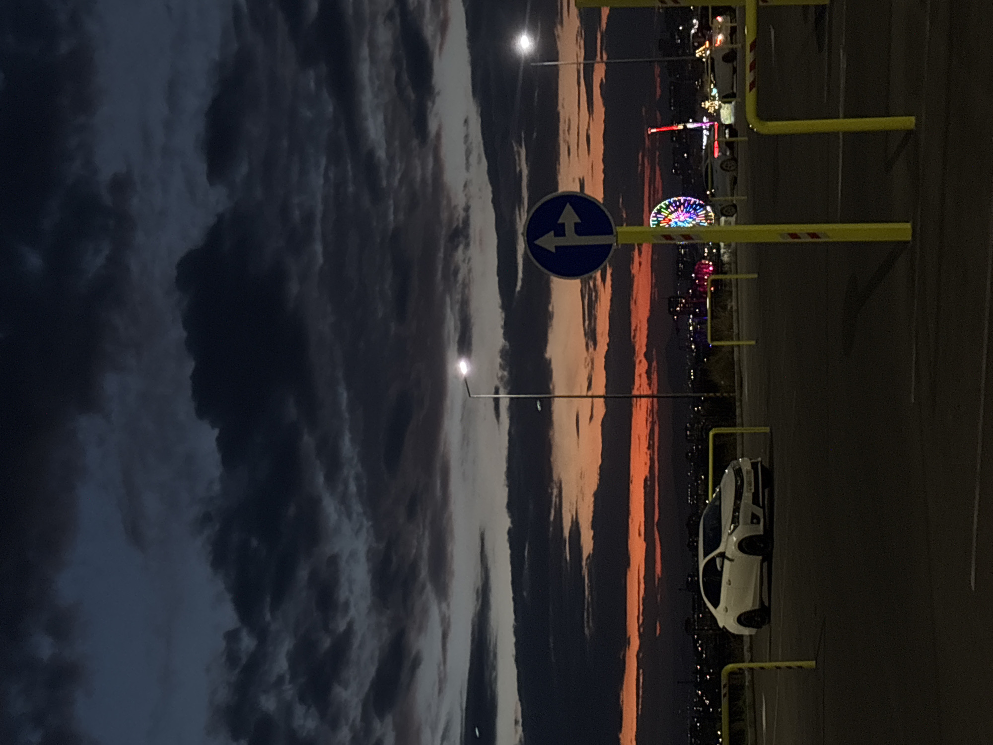 The image depicts a nighttime scene likely in Yerevan, Armenia. Dominating the left side are dark, overcast skies. The right side reveals an illuminated ferris wheel and other fairground attractions in the distance. A white car is parked in a parking space defined by yellow barrier poles. A blue and white traffic sign indicating left or right is also visible. The background shows city lights. The scene is lit by streetlights and the illuminations from the fairground.