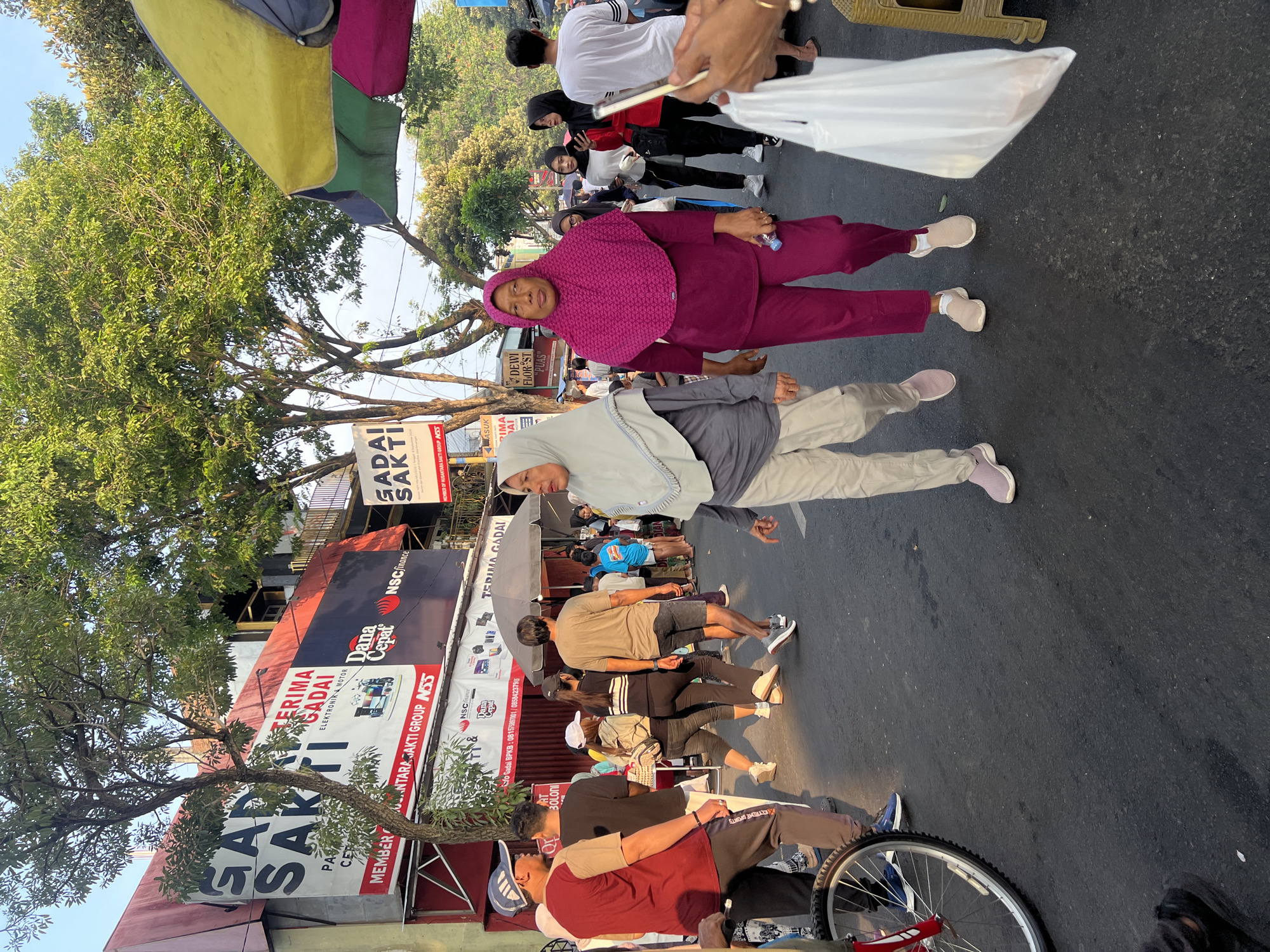 The image captures a street scene in Kecamatan Pati, Indonesia, likely a market or bustling commercial area given the presence of stalls and diverse pedestrians. In the foreground, two women walk towards the viewer. The woman on the left wears a light grey hijab and a grey long-sleeved top with light beige pants and pink athletic shoes. The woman on the right is dressed in a bright fuchsia hijab and matching fuchsia pantsuit with beige slip-on shoes. She is holding a water bottle.
Behind them, a crowd of people are seen walking, shopping, and standing. A man wearing a maroon t-shirt and a blue cap is visible on the left, facing away from the camera, with a bicycle wheel partially in frame. Other individuals in the background are wearing casual clothing, including t-shirts, pants, and hijabs, suggesting a mixed demographic. Some people are carrying bags.
The environment includes buildings with signage, the largest being a prominent banner that reads "GADAI SAKTI" in large black letters, with smaller text below indicating services like "TERIMA GADAI" (Pawn Accepted) and business names like "DANA CEPAT" and "NSC FINANCE". Another sign visible further down the street also displays "GADAI SAKTI". Several trees with dense green foliage provide shade, and colorful umbrellas are set up, one predominantly yellow and green, another appearing pink or purple. The street is paved with asphalt, and the time of day appears to be late afternoon due to the warm lighting and shadows cast. The overall atmosphere suggests a lively commercial district with active street life.