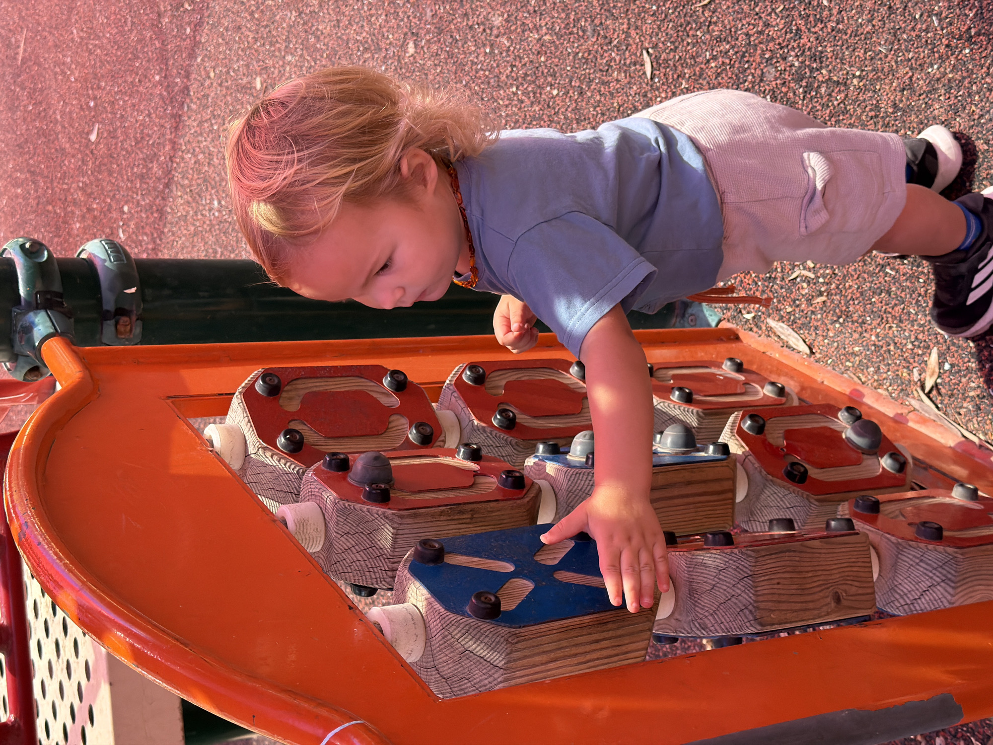 A child with blonde hair, wearing a blue shirt, beige shorts, and black and white athletic shoes, is playing on an orange and wooden playground structure. The structure features various wooden blocks with colorful tops (red and blue) and black knobs, designed for tactile exploration. The playground surface around the structure appears to be composed of small gravel. The image was taken in Kiryat Motzkin, Israel.