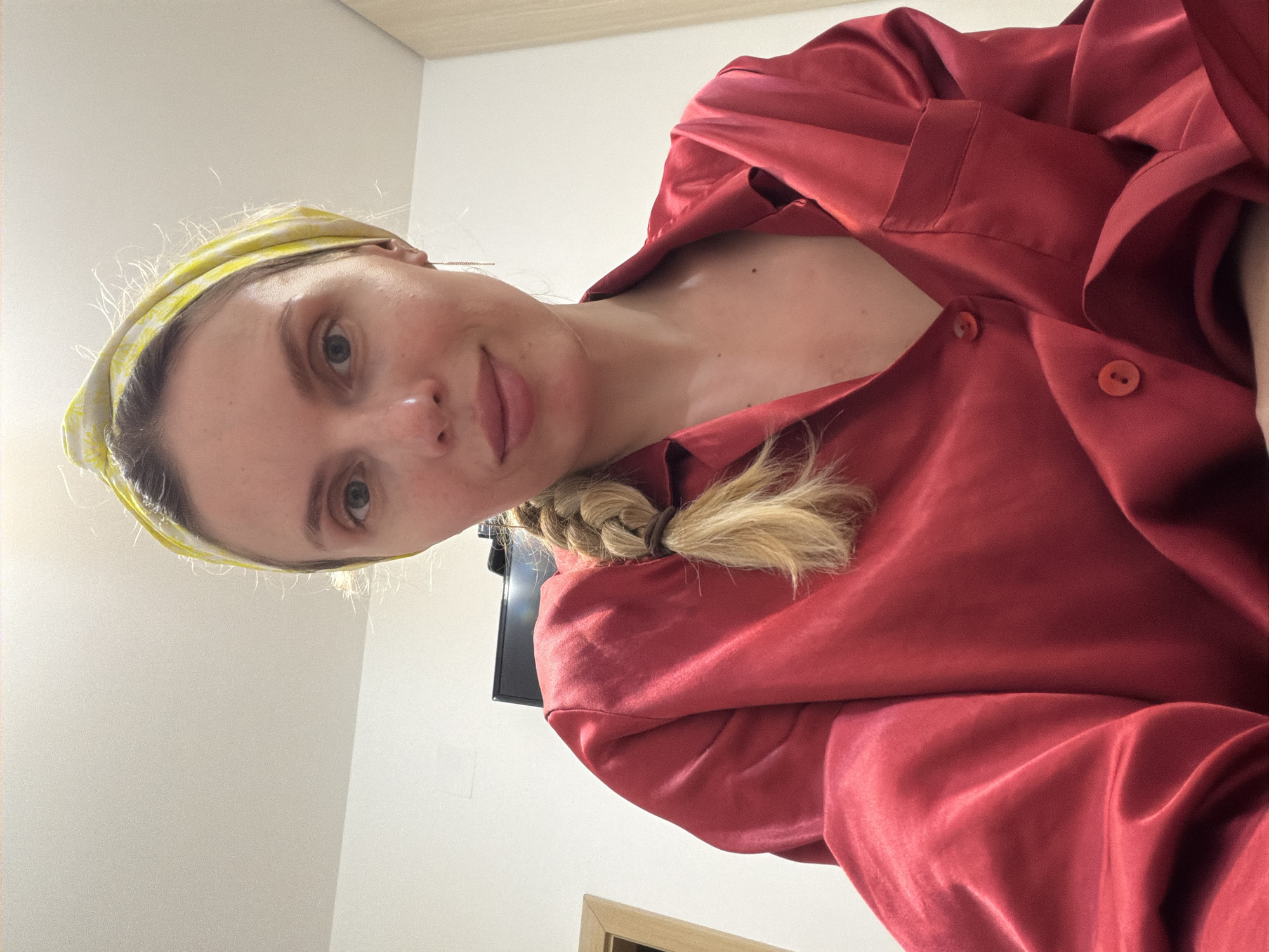 A woman with blonde hair and a yellow headband is pictured wearing a red shirt. She is looking at the camera. There is a TV screen behind her on the wall. A framed picture is partially visible in the background. The location is Budva, Montenegro.